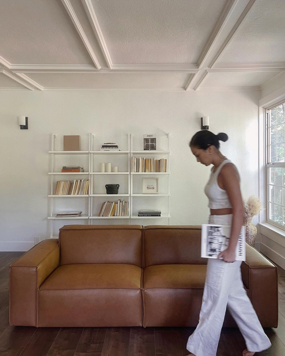 A person holding a magazine walking past a leather sofa.