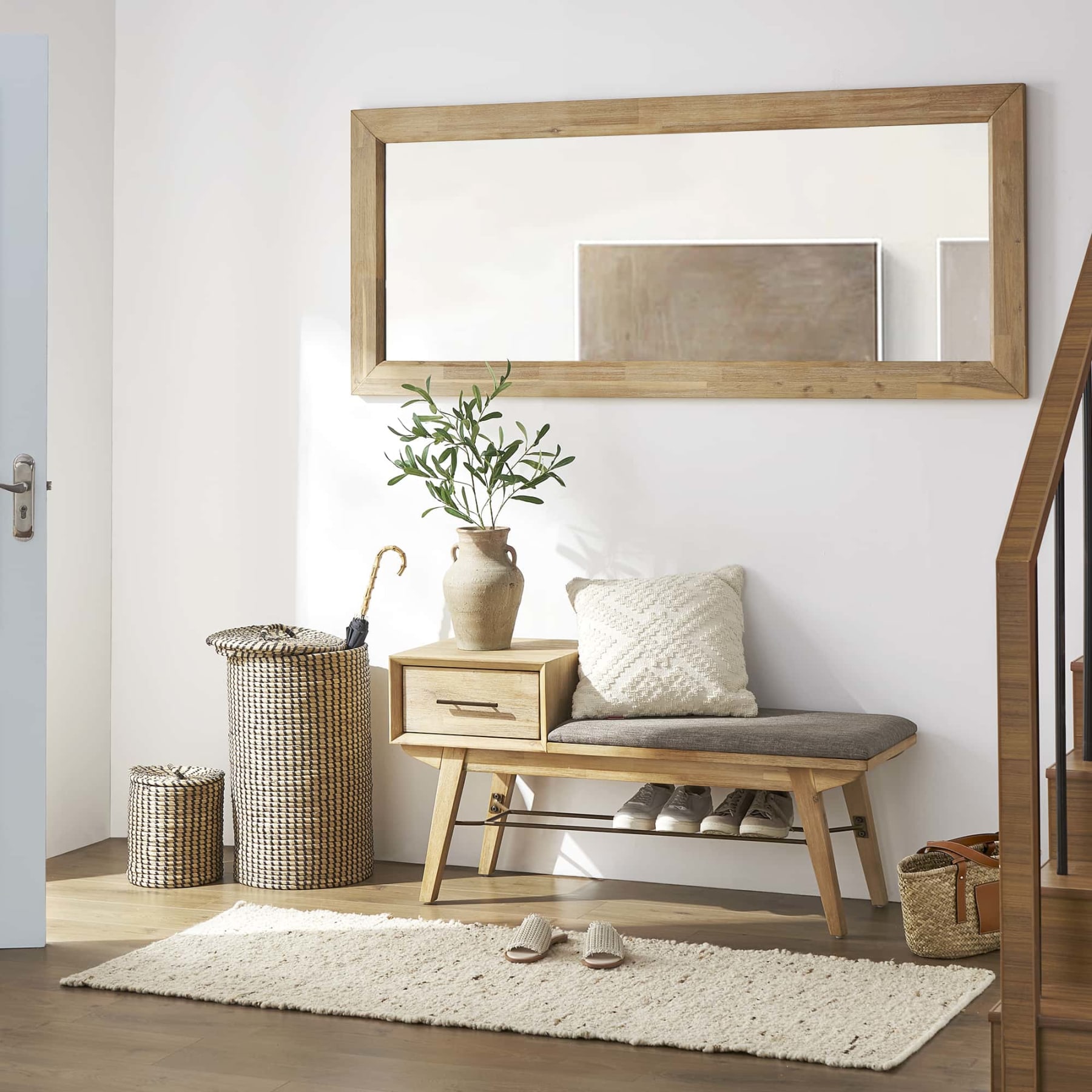 A light wood entryway bench with storage and shoes tucked below it along with a wooden frame mirror and white wool runner rug