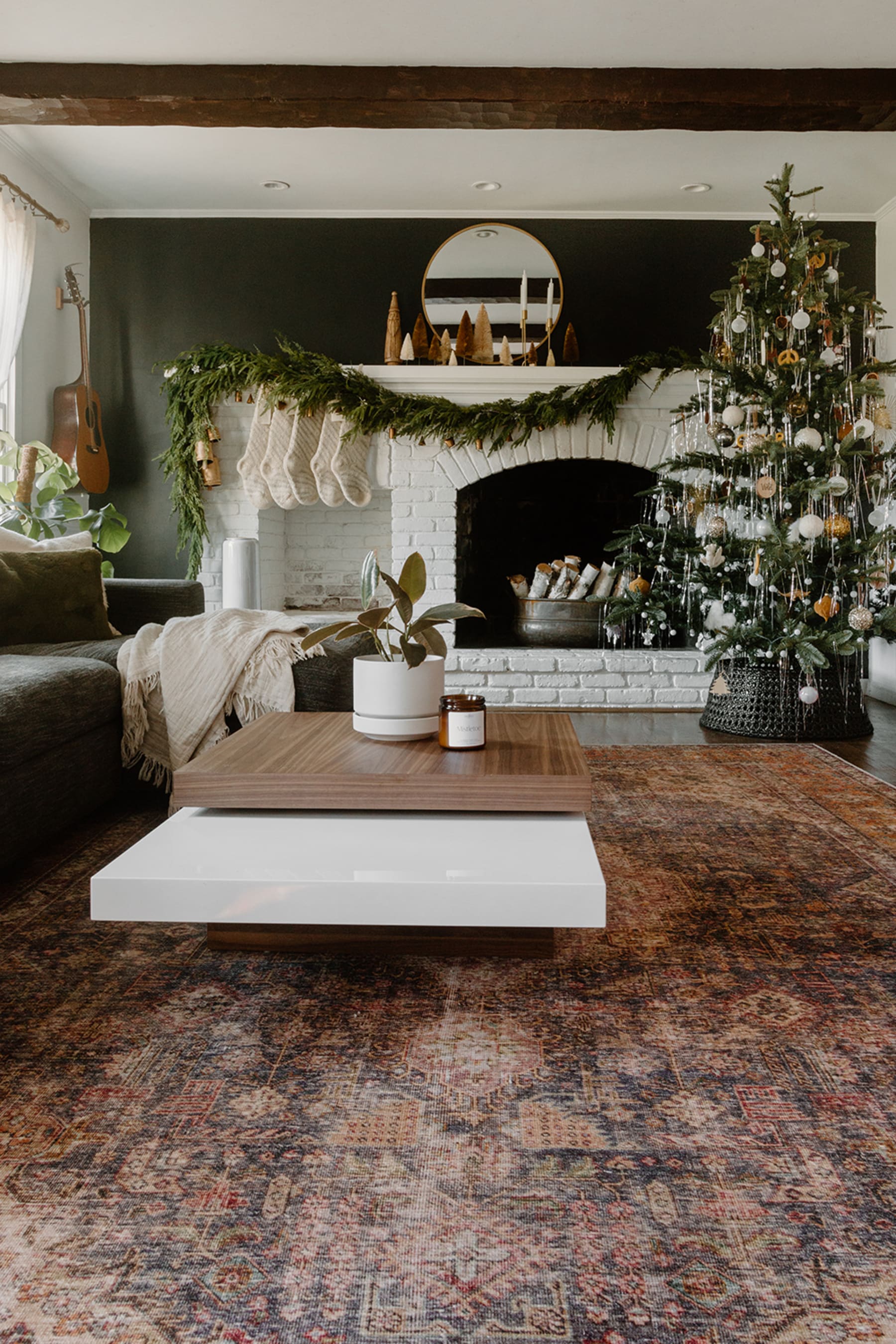 A designer coffee table beside a fireplace with a potted plant on a rug in a dark living room with Christmas decorations and a long area rug.