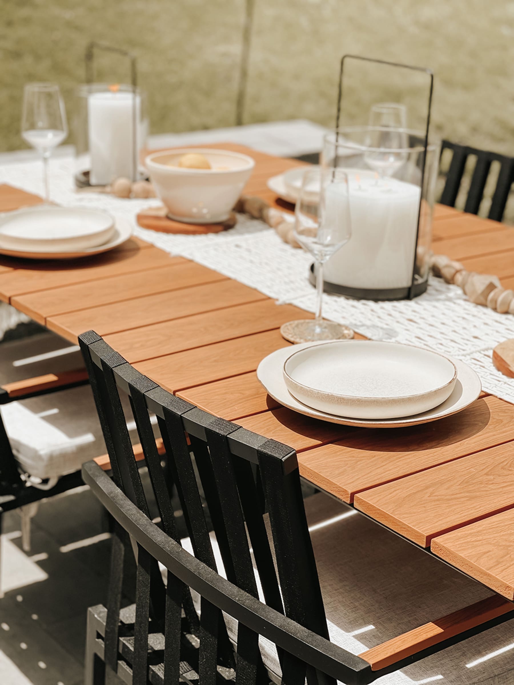 A wooden outdoor dining table set up for an outdoor dining session with porcelain plates and cutlery decorated with a candle and a cloth