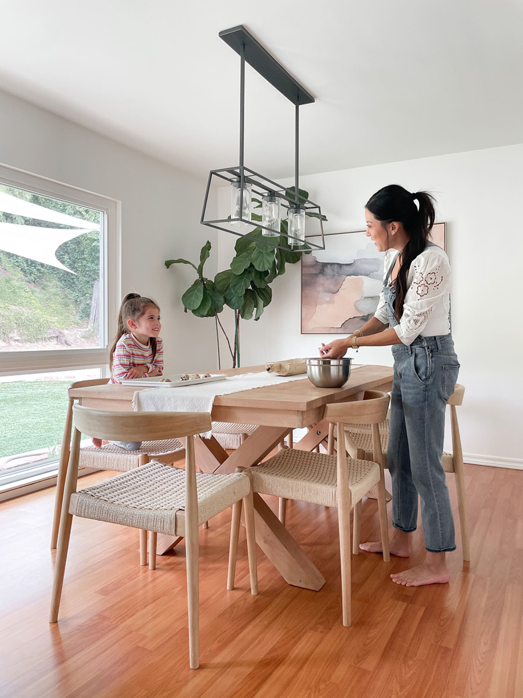 A young girl watches her mother clean and prepare a wooden table before a meal in the dining room against a transparent glass door and tall potted plant