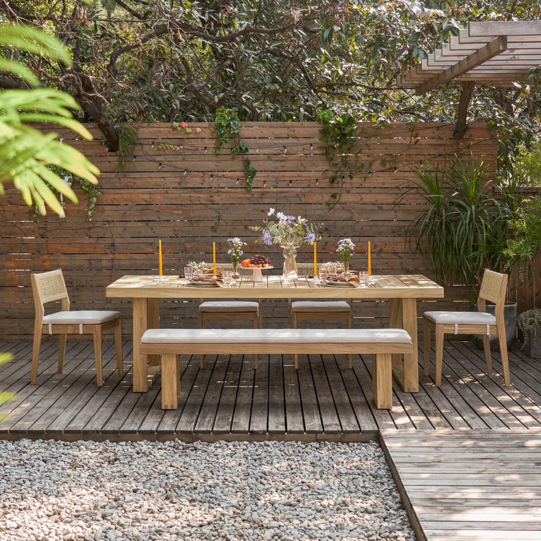 A teak dining table with a bench and 4 chairs sit on a wooden deck.