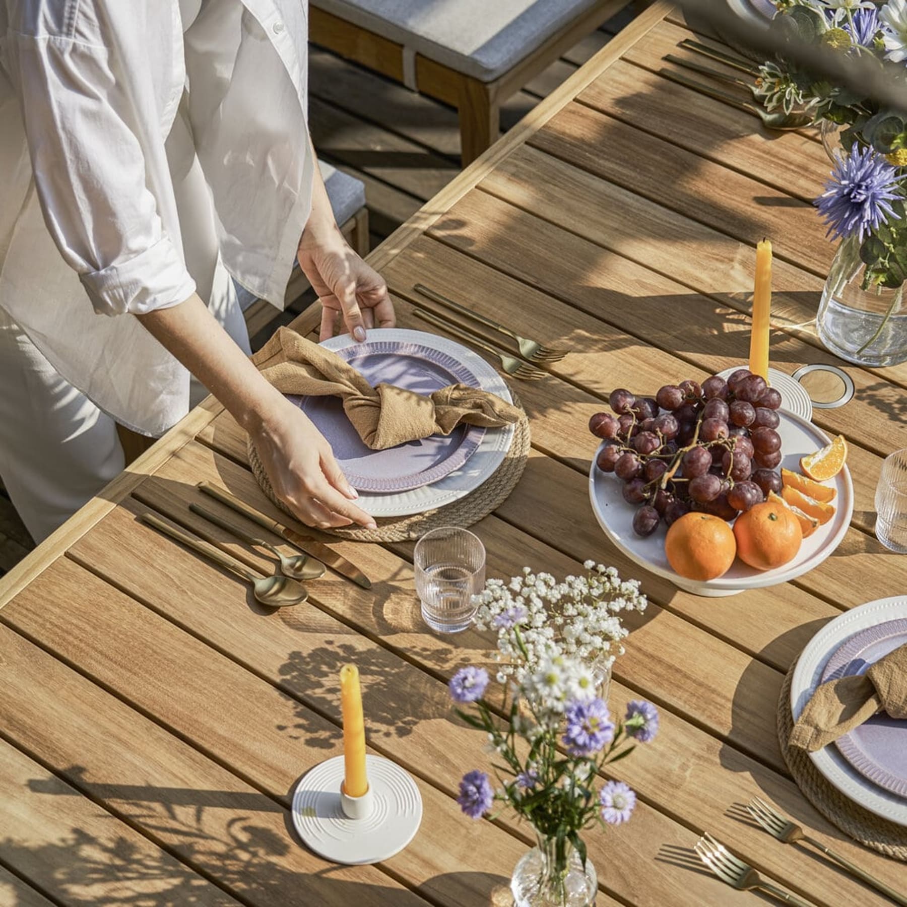 Dishes and cups are placed on an outdoor teak dining table with woven placemats.