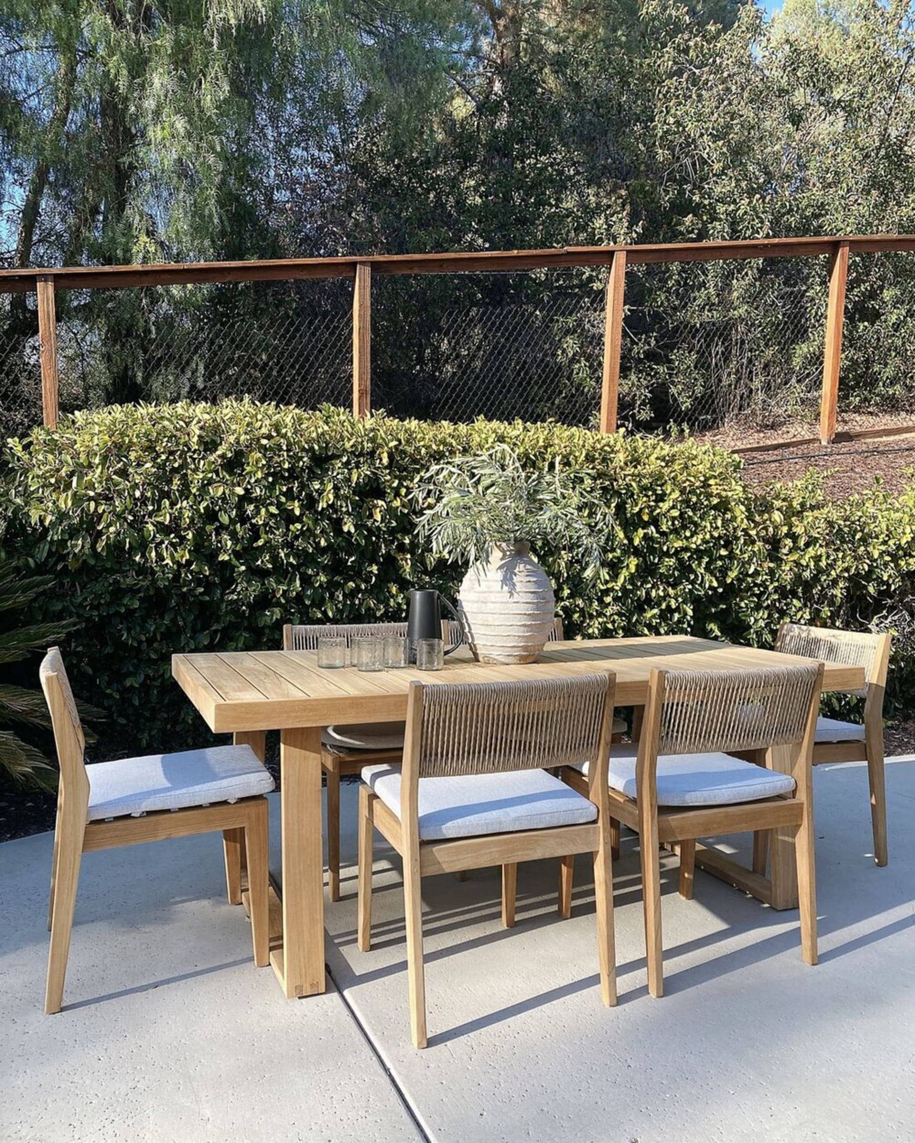 A teak outdoor dining table with a vase, water kettle, and glasses atop.