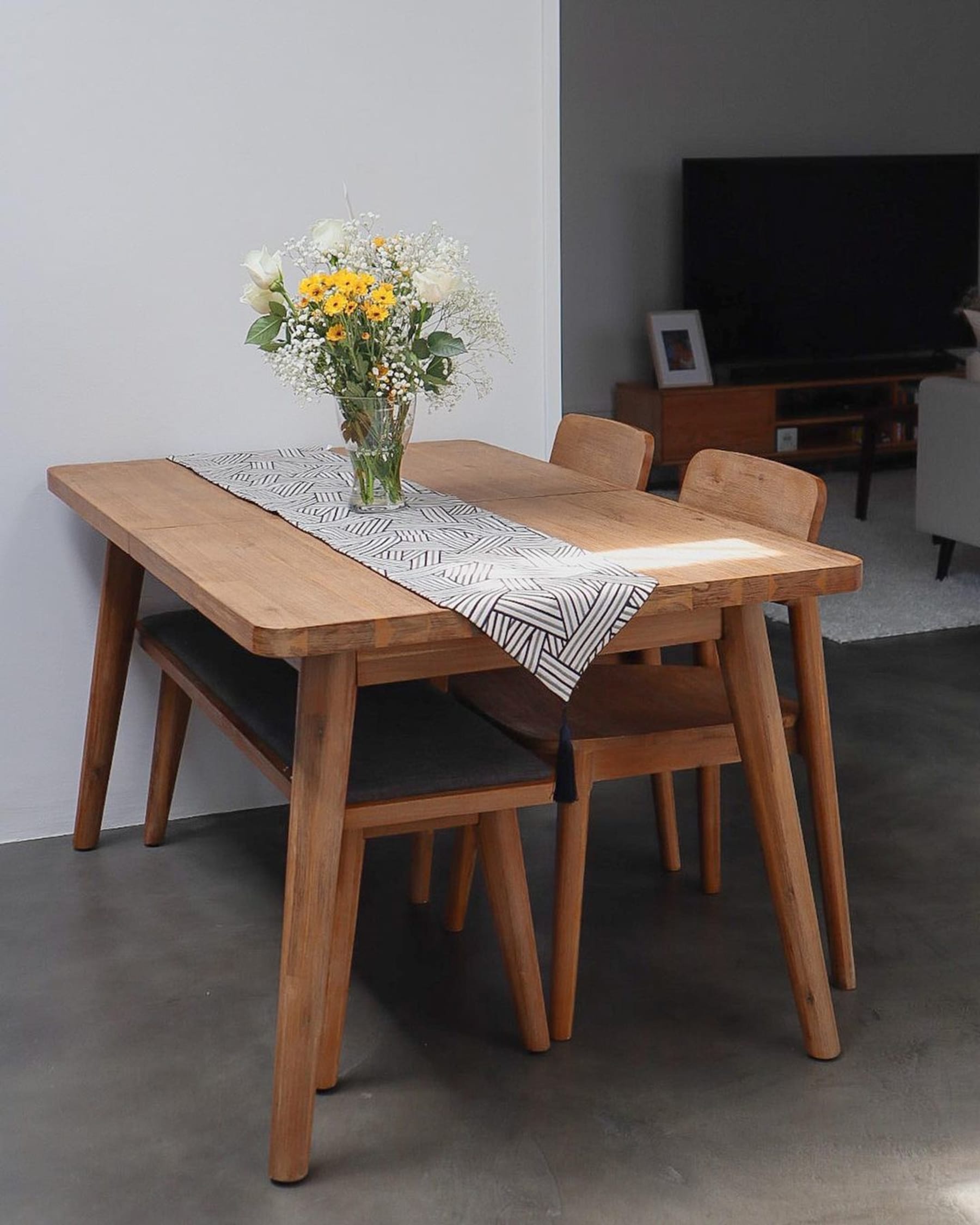 A wooden extendable dining table set with 2 chairs and a bench has an abstract runner and a vase of flowers.