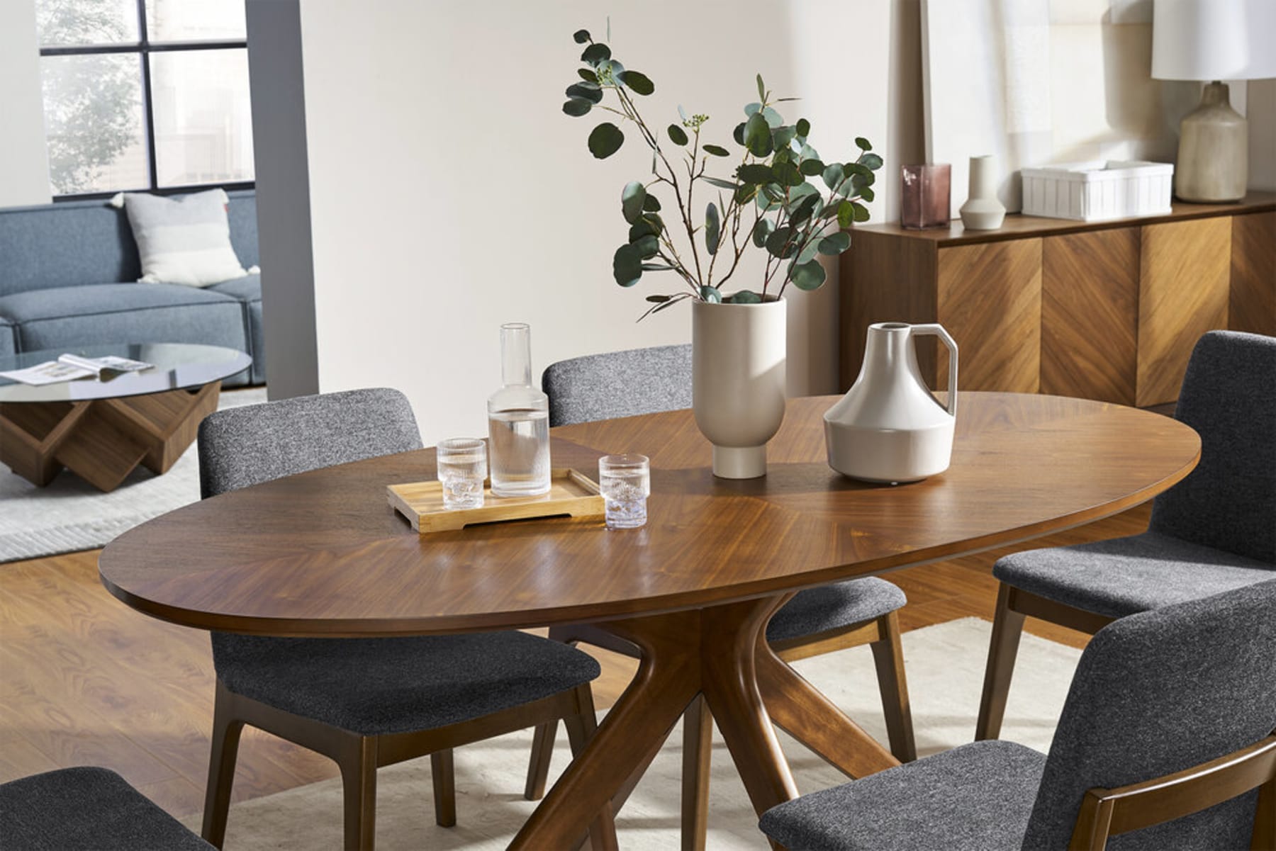 A wooden oval dining table is decorated with a vase, a tray, and a jug.