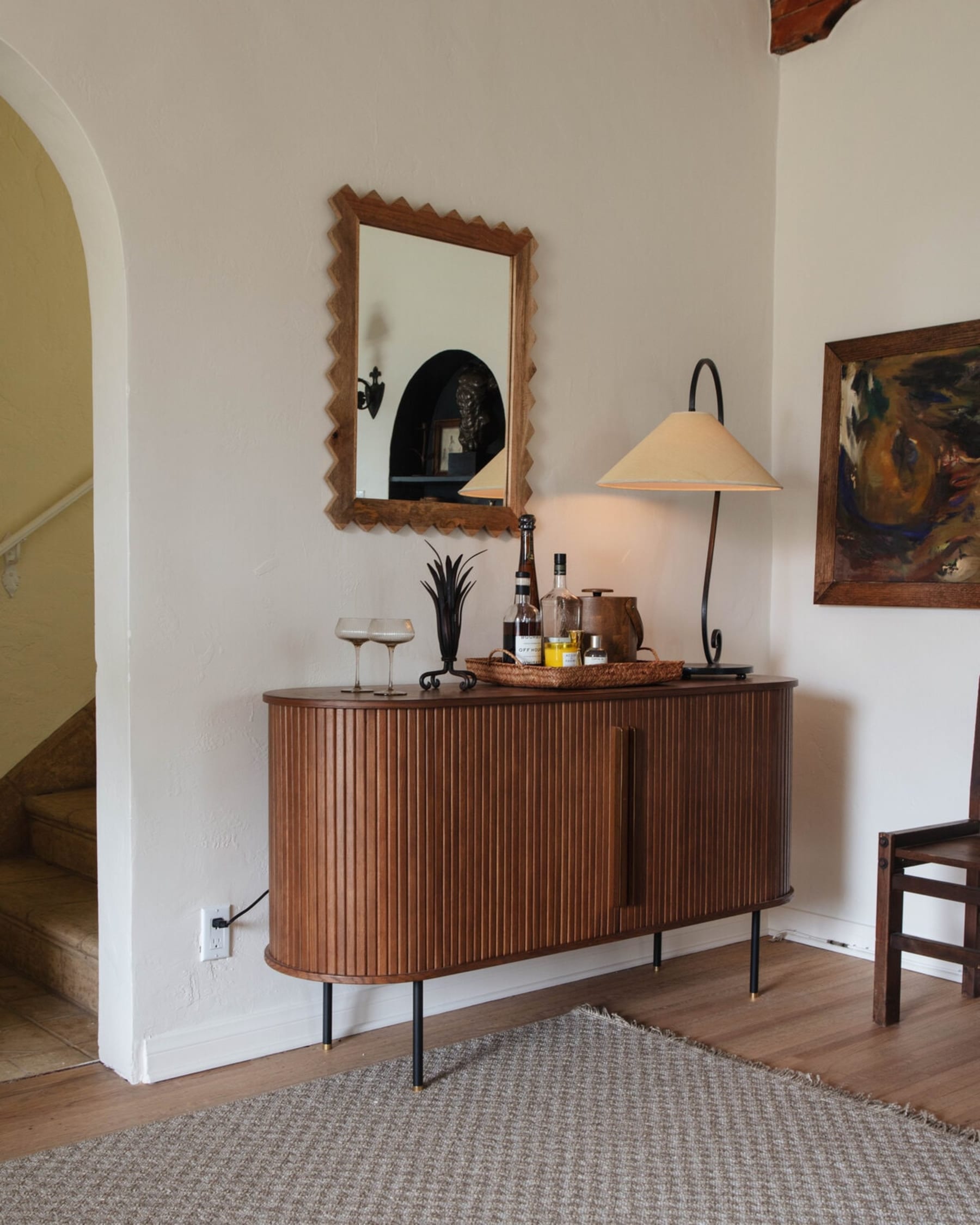 A wooden sideboard with an oval silhouette has a lamp, wine glasses, and some drinks on top.