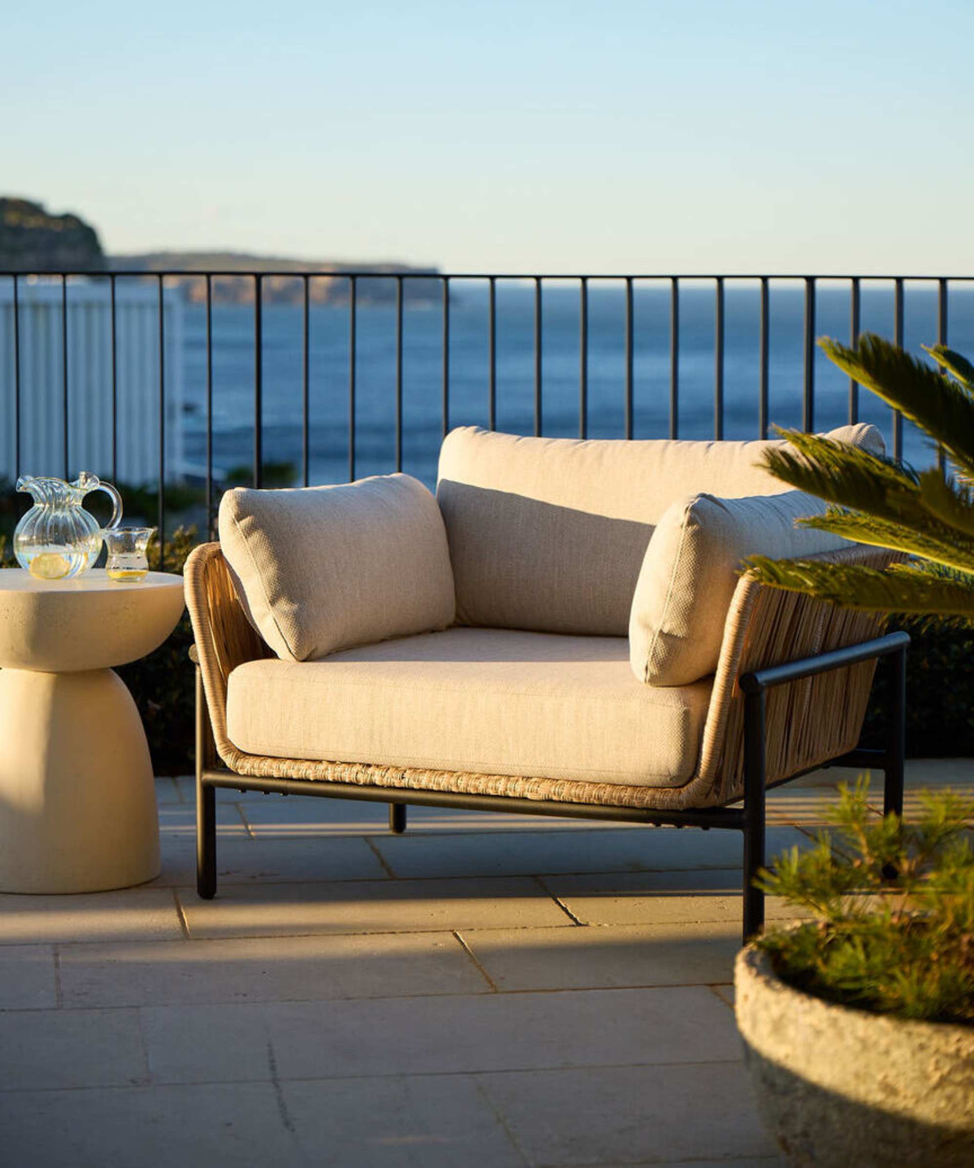 An outdoor wicker lounge chair with a metal frame placed on a balcony overlooking the ocean.