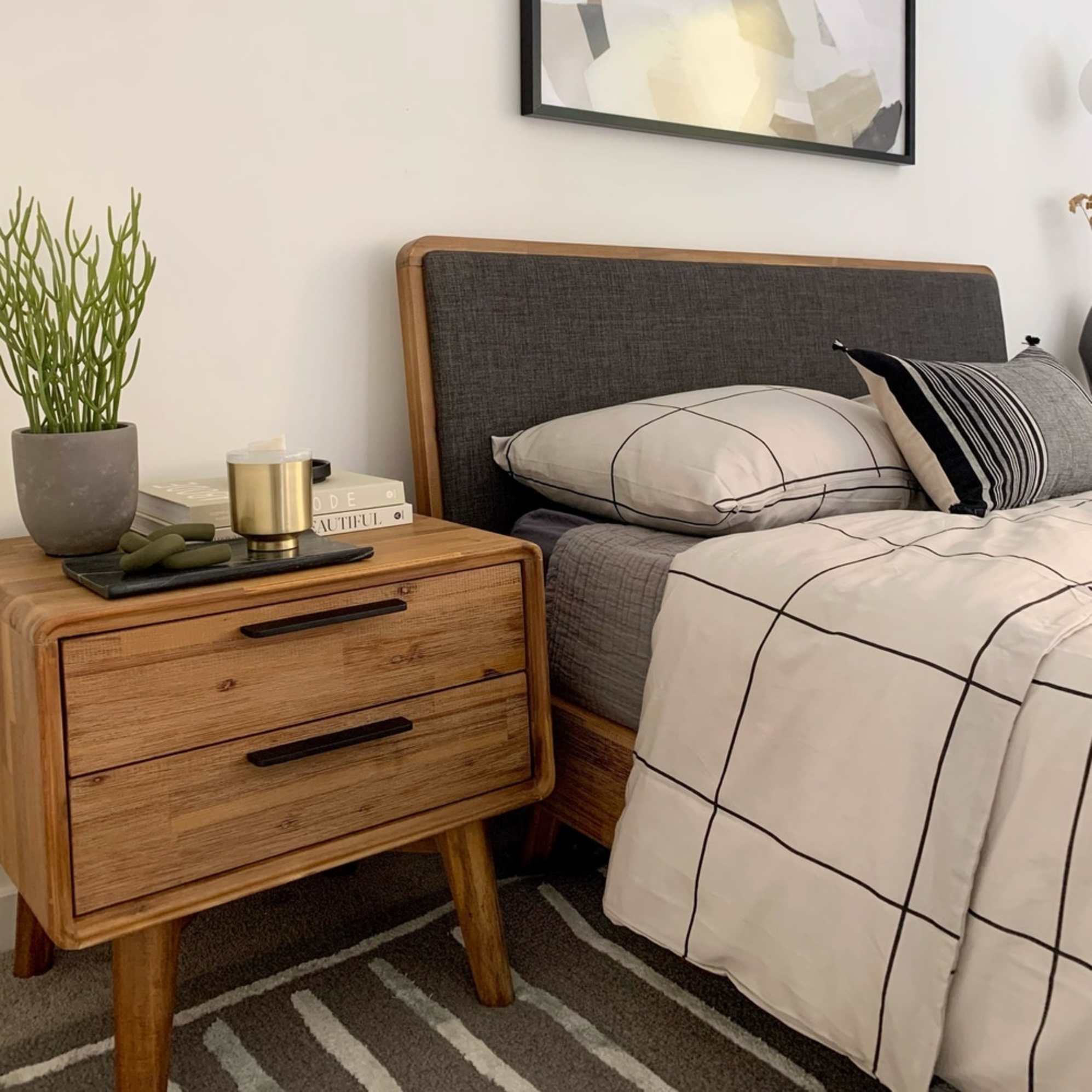An acacia wood with a fabric headboard and a matching bedside table decorated with a plant and books.