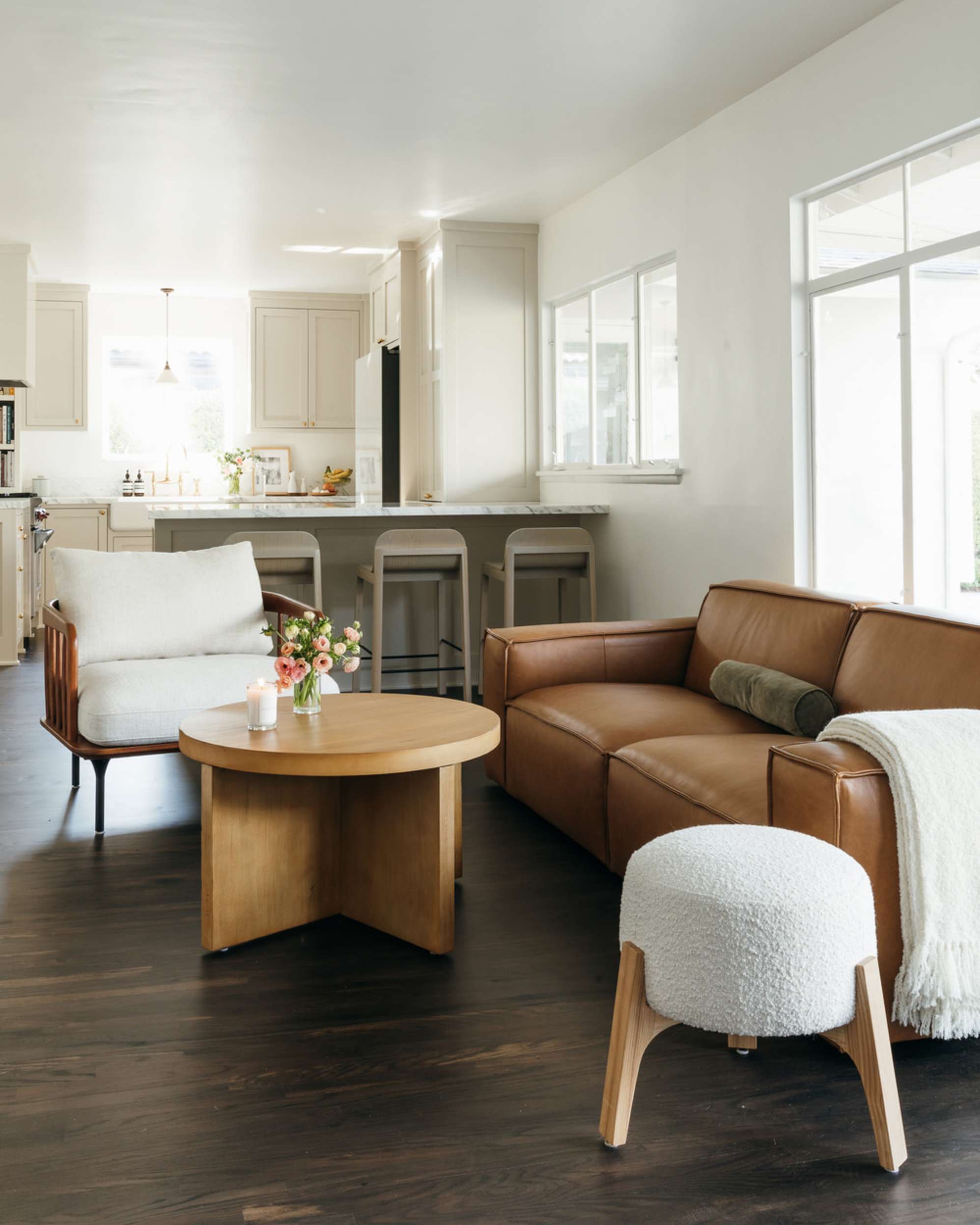 A leather sofa, round wooden coffee table, bouclé stool, and wooden armchair in a living room.