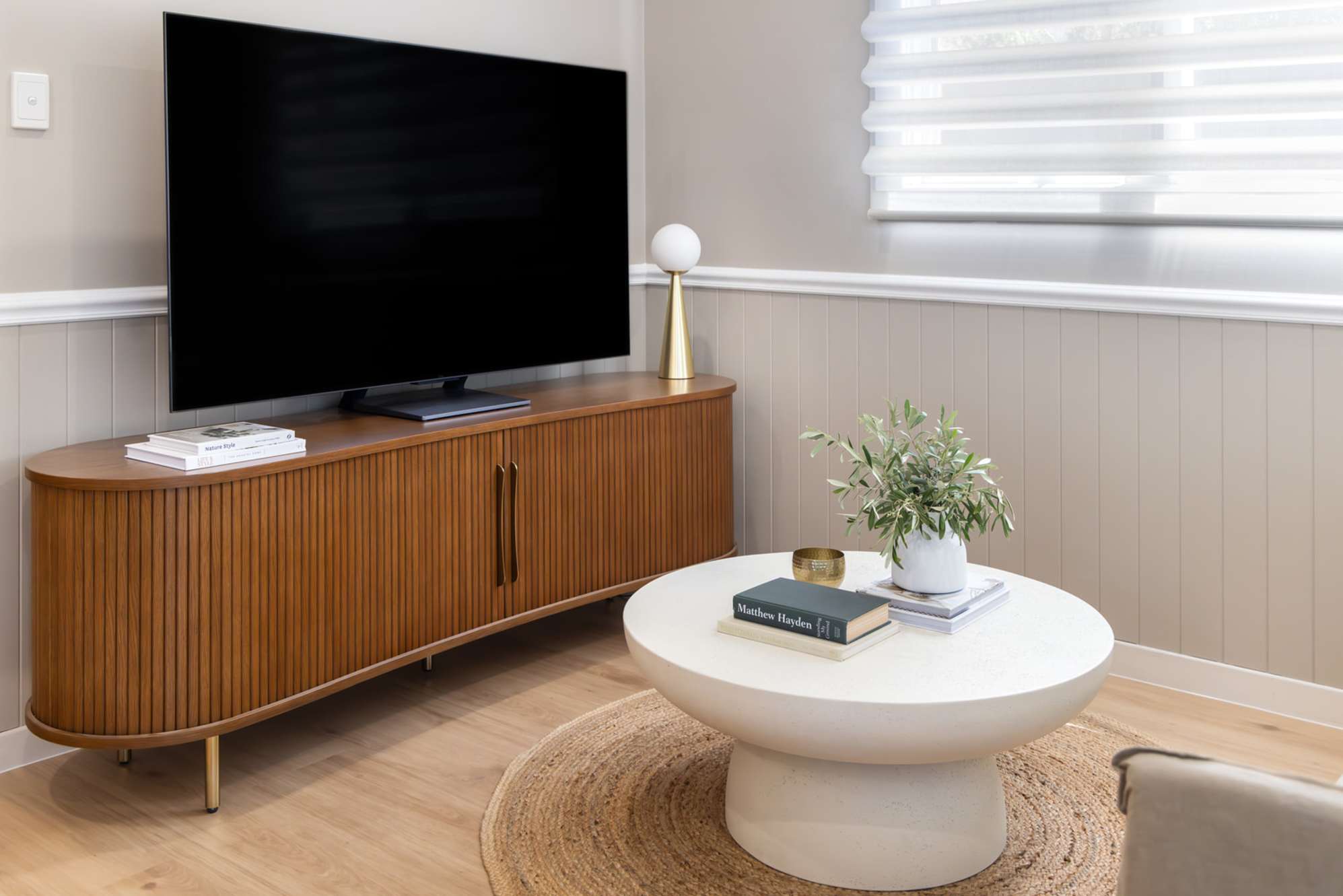 A wooden curved TV stand placed in a living room with a round white coffee table.