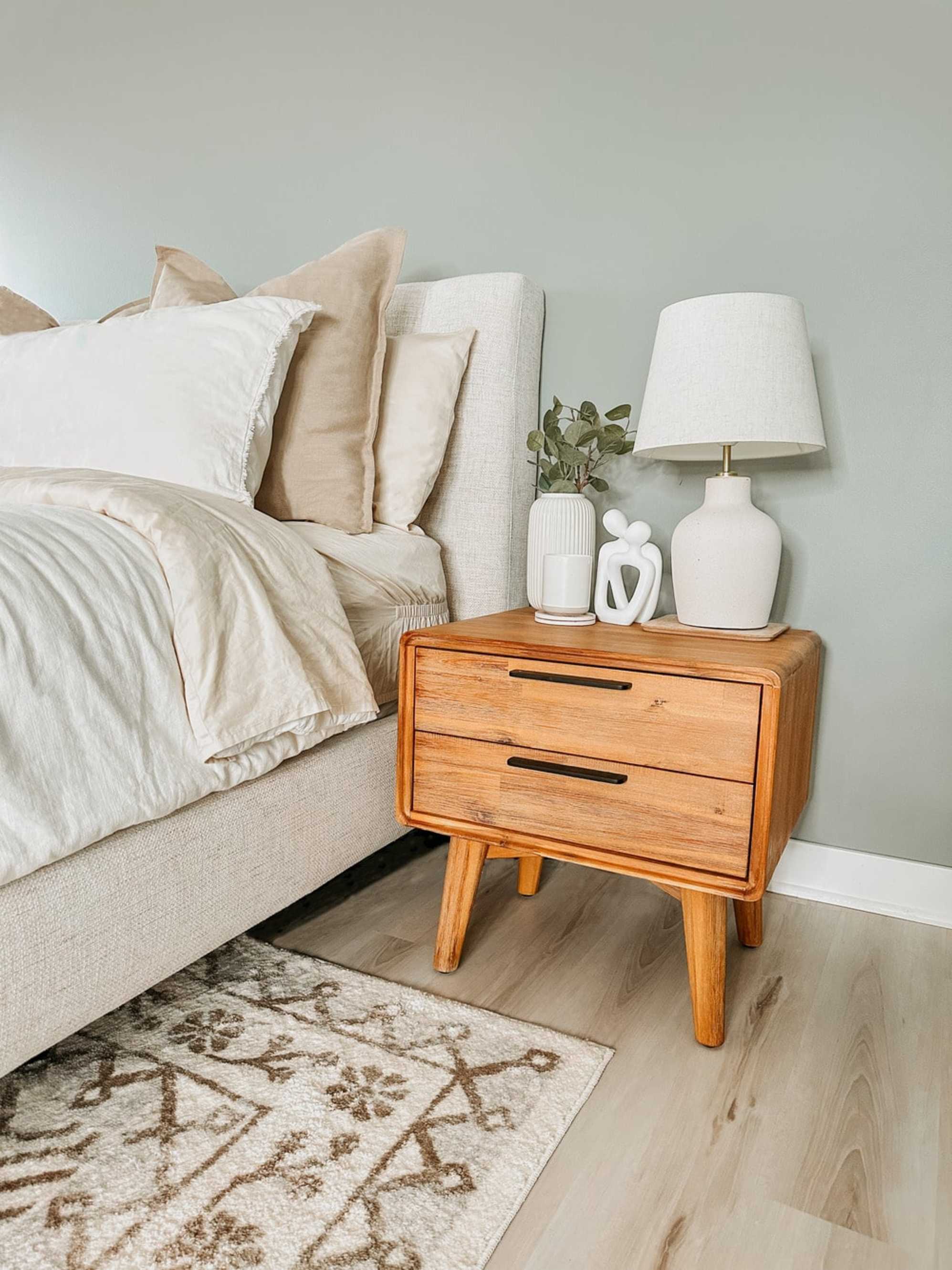 An acacia wood nightstand with a vase, lamp, and decor.