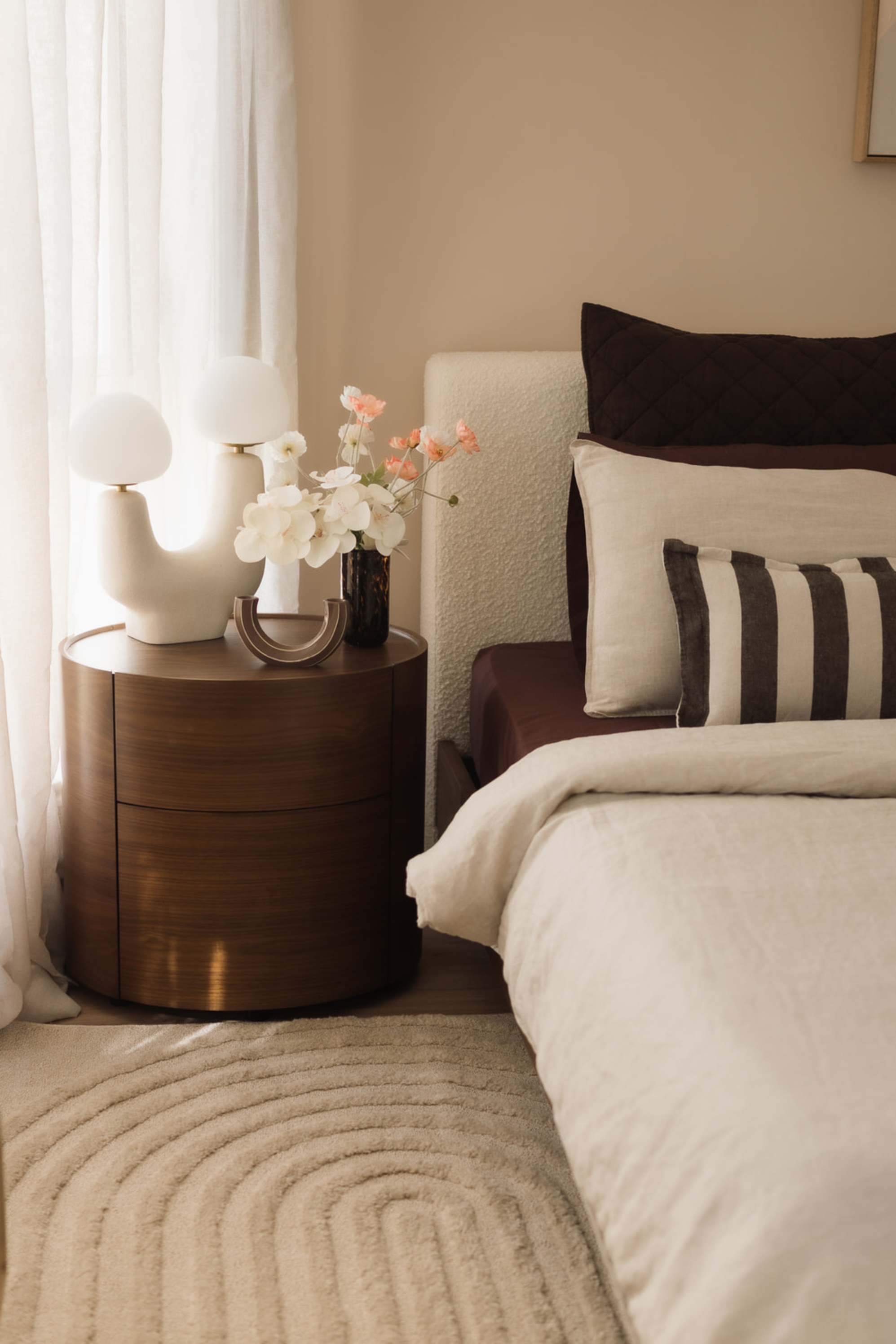 A wooden round side table with a sculptural lamp atop placed beside a bed.