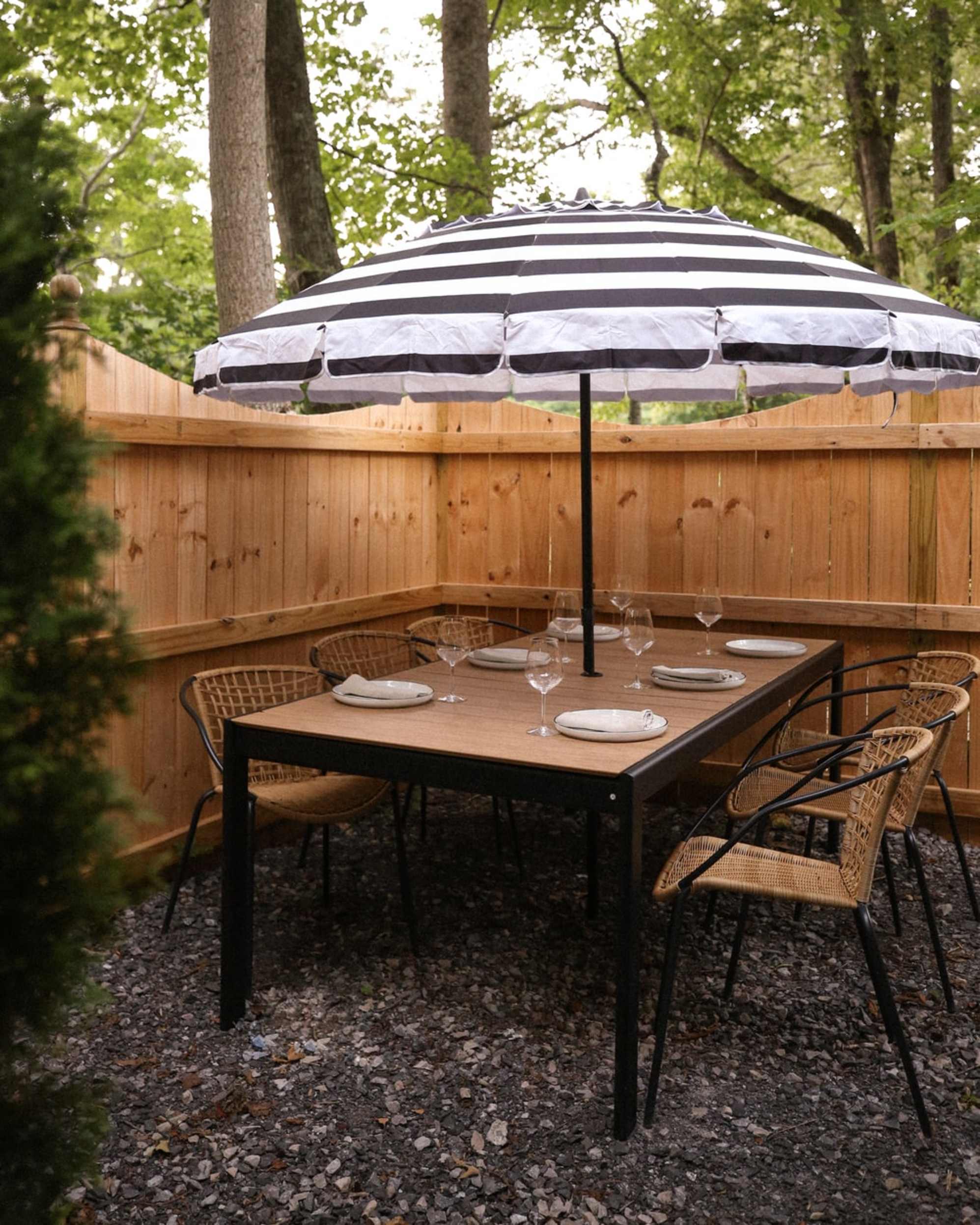An outdoor dining table and six matching chairs are placed in a backyard with an outdoor umbrella.