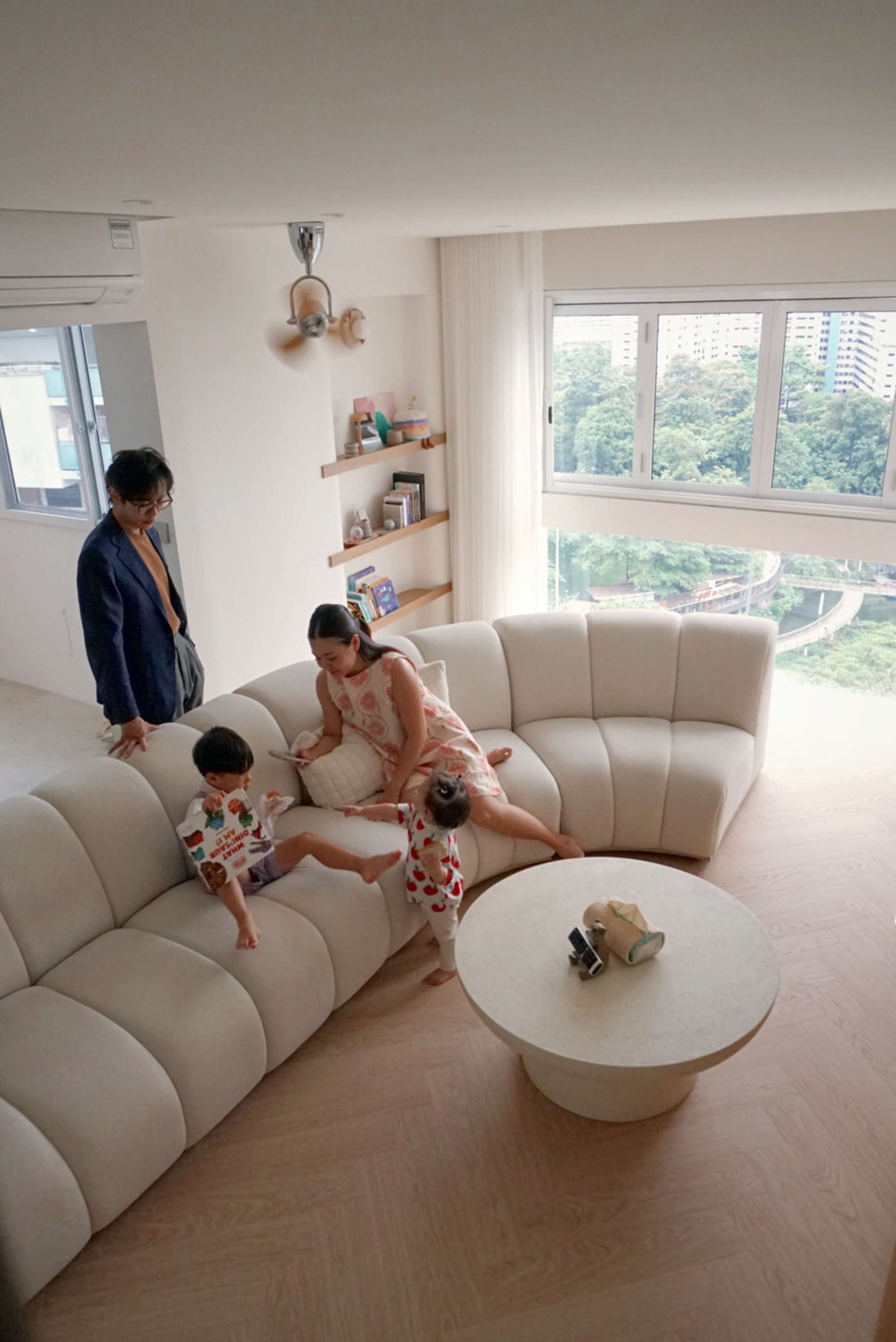 Two people and two toddlers playing around a curved sofa and a round coffee table that are placed in a living room.