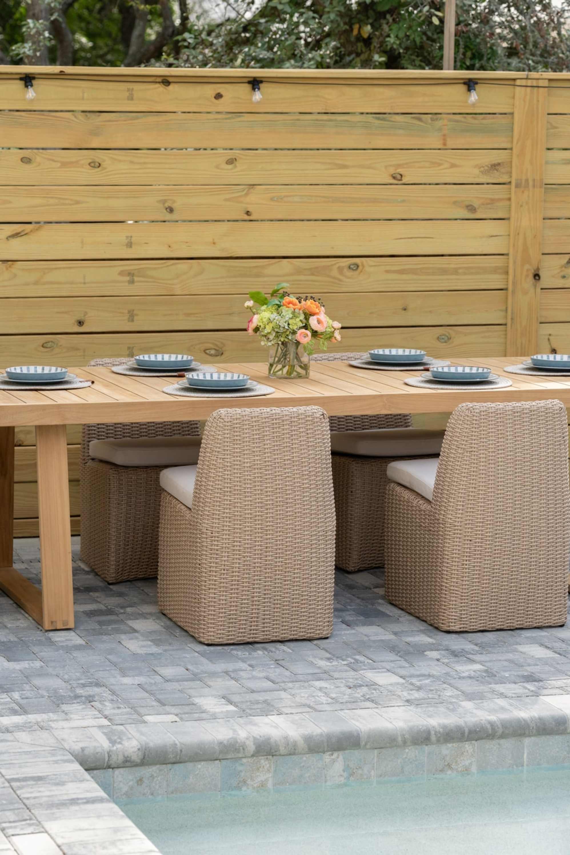 A teak outdoor dining table and 4 wicker chairs are placed by a pool.
