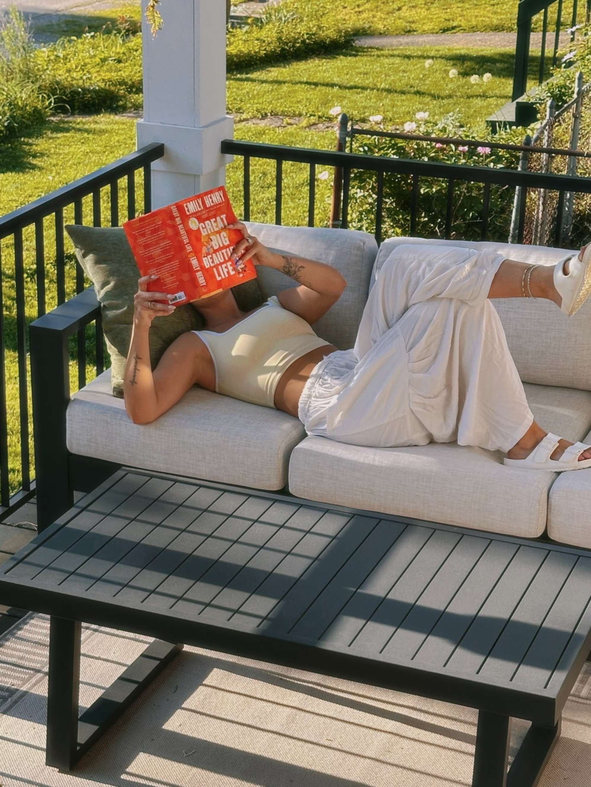 A woman lying down on an outdoor aluminum sofa while reading a book.