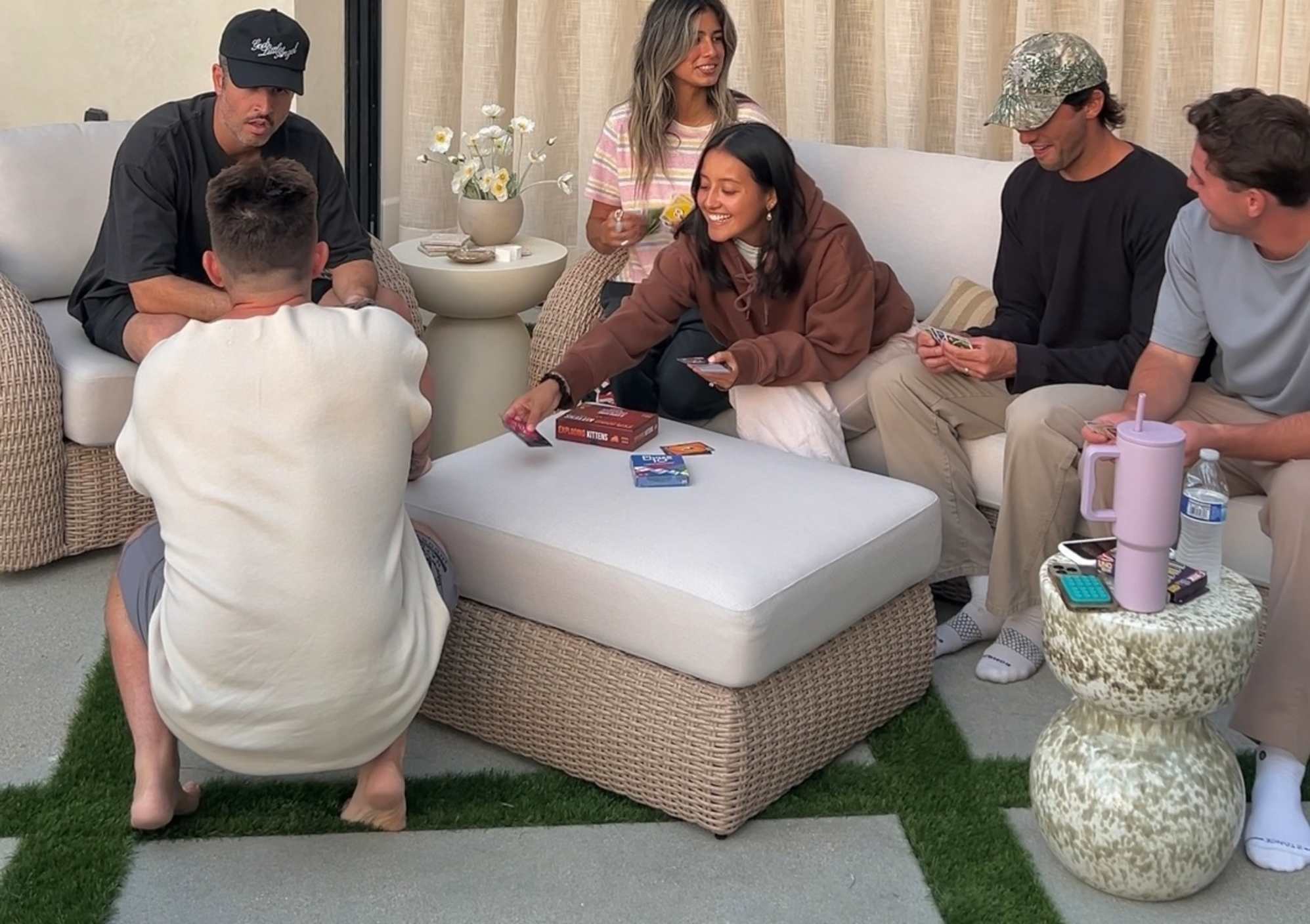 A group of people seated on an outdoor sofa while playing card games on the outdoor ottoman-coffee table.
