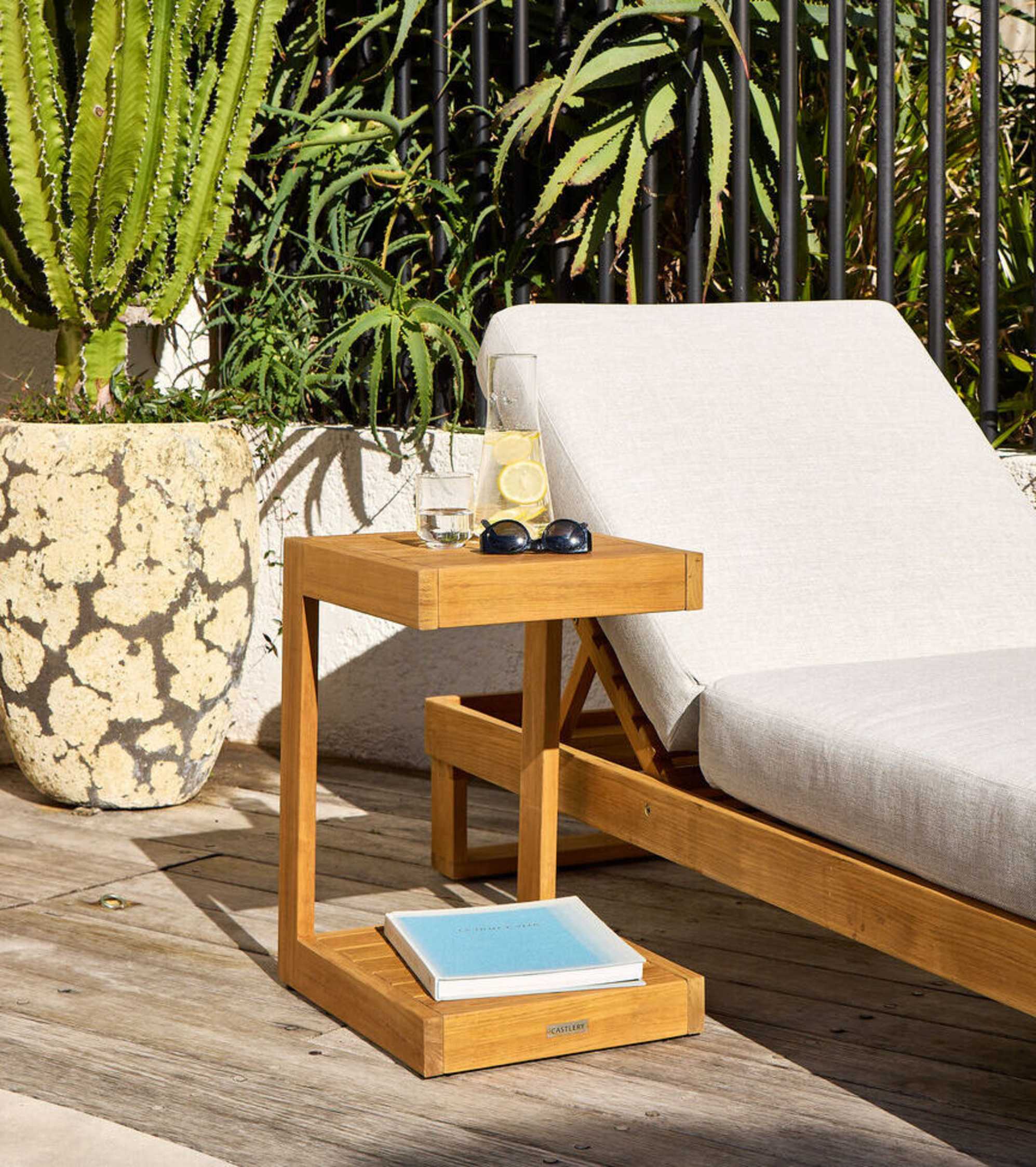 A teak outdoor c-side table with a jug of water, sunglasses, and a book placed beside a matching teak lounger.