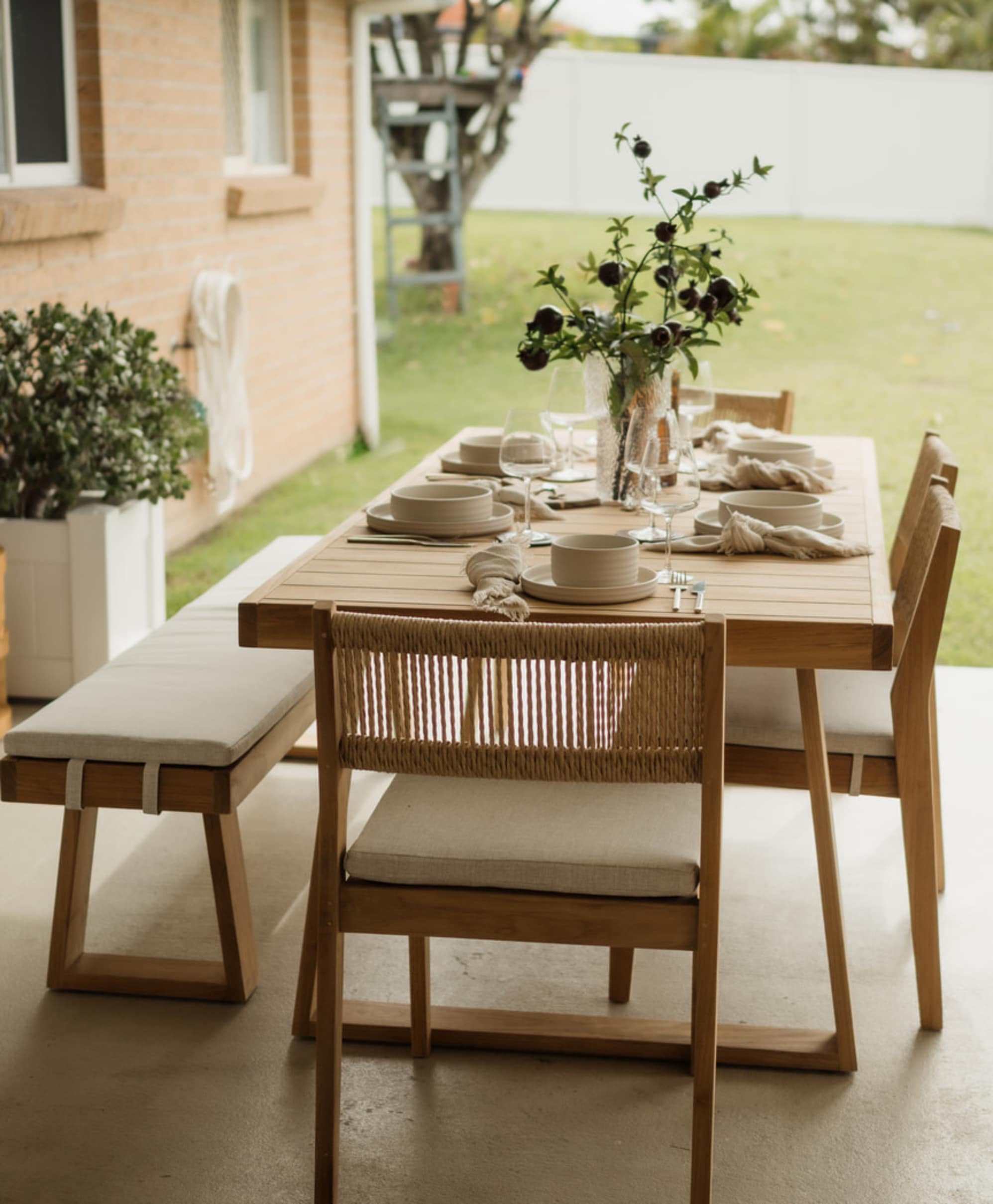 An outdoor teak dining table set lined with white tableware and a vase.