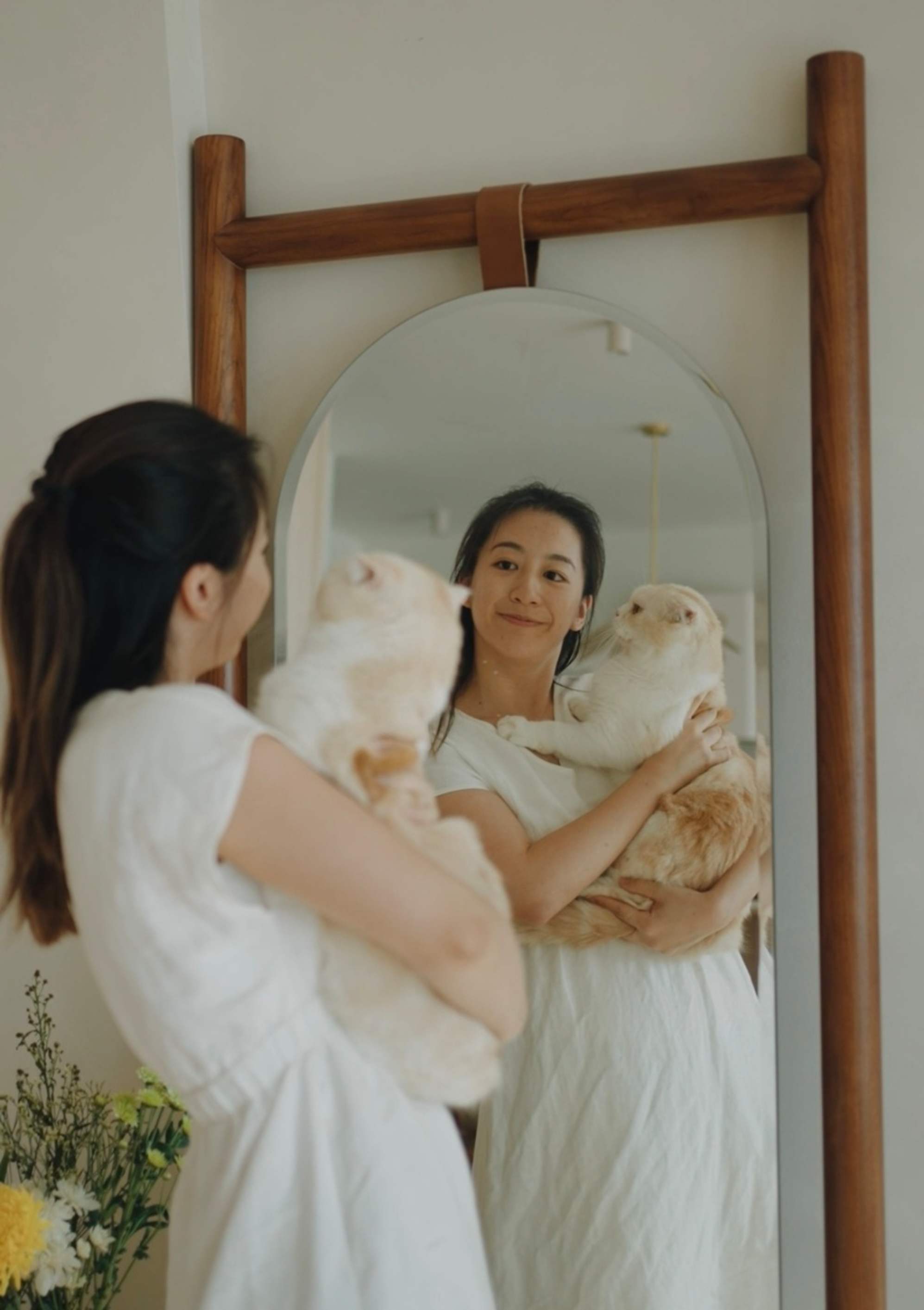 A person holding her cat while smiling at a floor mirror propped up against a wall at the entrance.