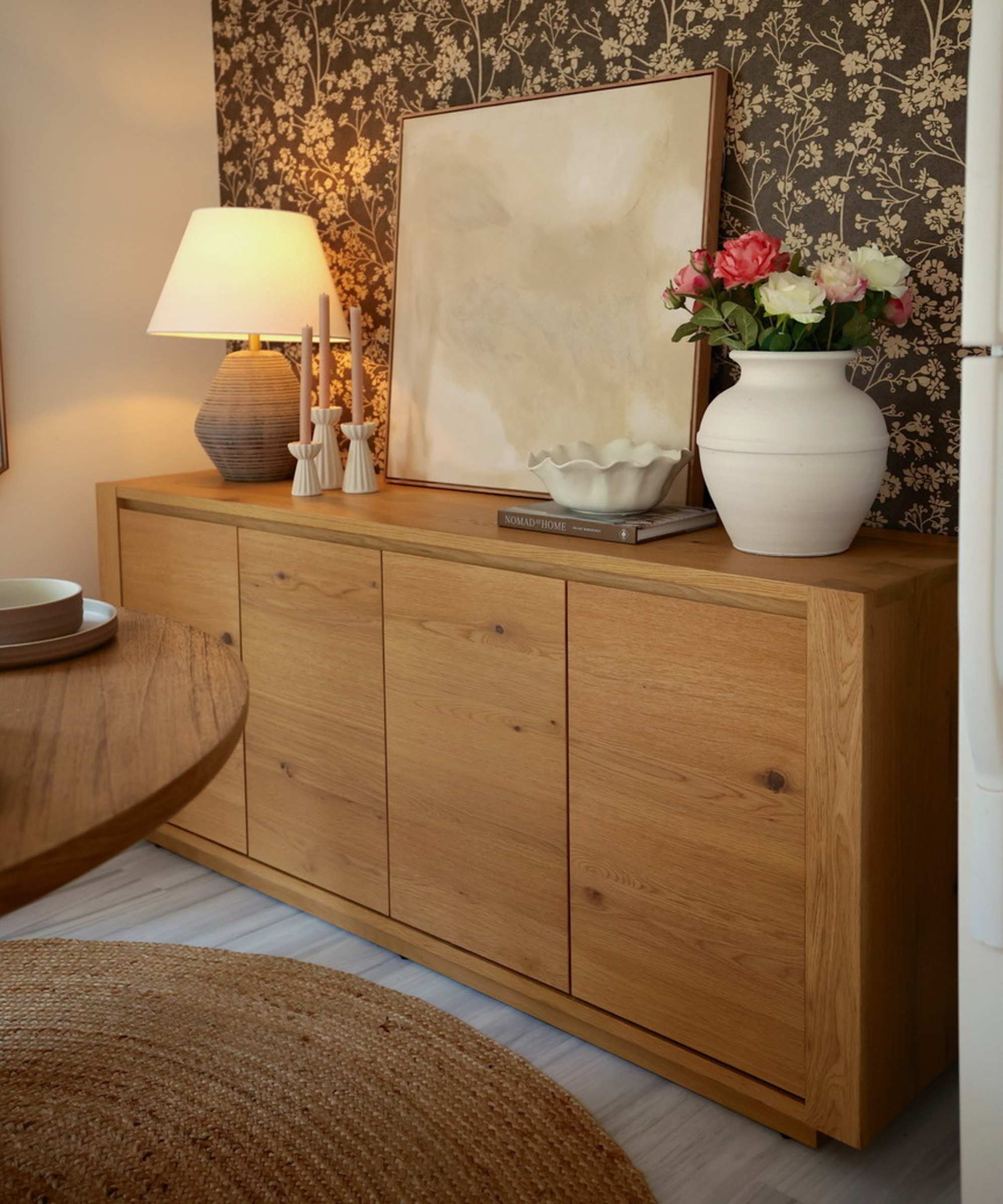 A wooden sideboard placed in a dining area against an accent wall with a vase, lamp, and artwork.
