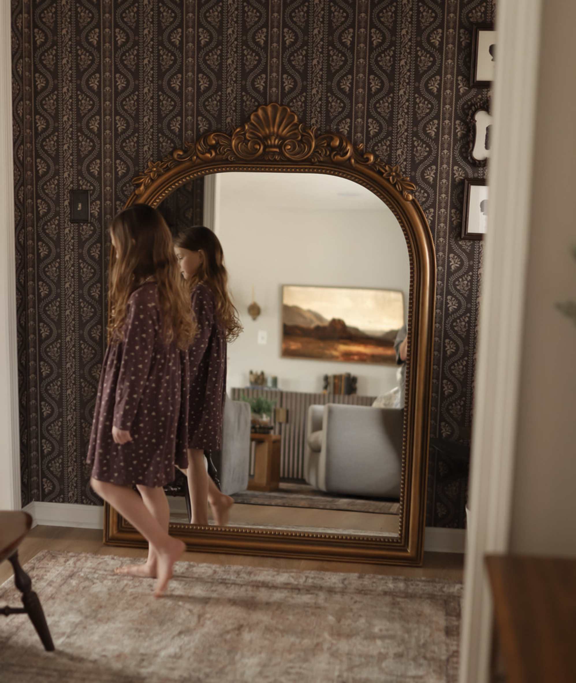 A little girl wearing a brown dress walking infront of a floor mirror propped up against a wall.