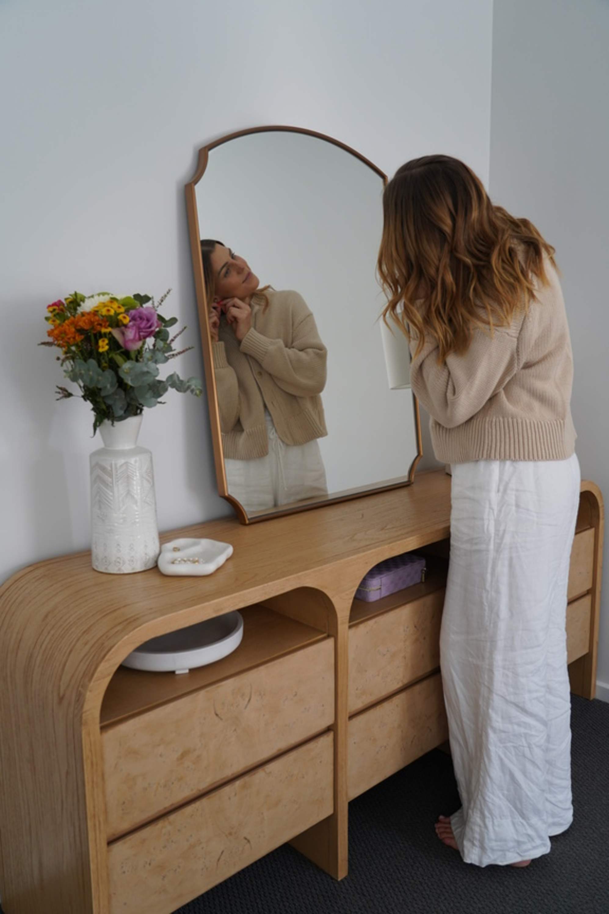  A woman standing infront of a dresser with a mirror while fixing her earrings.