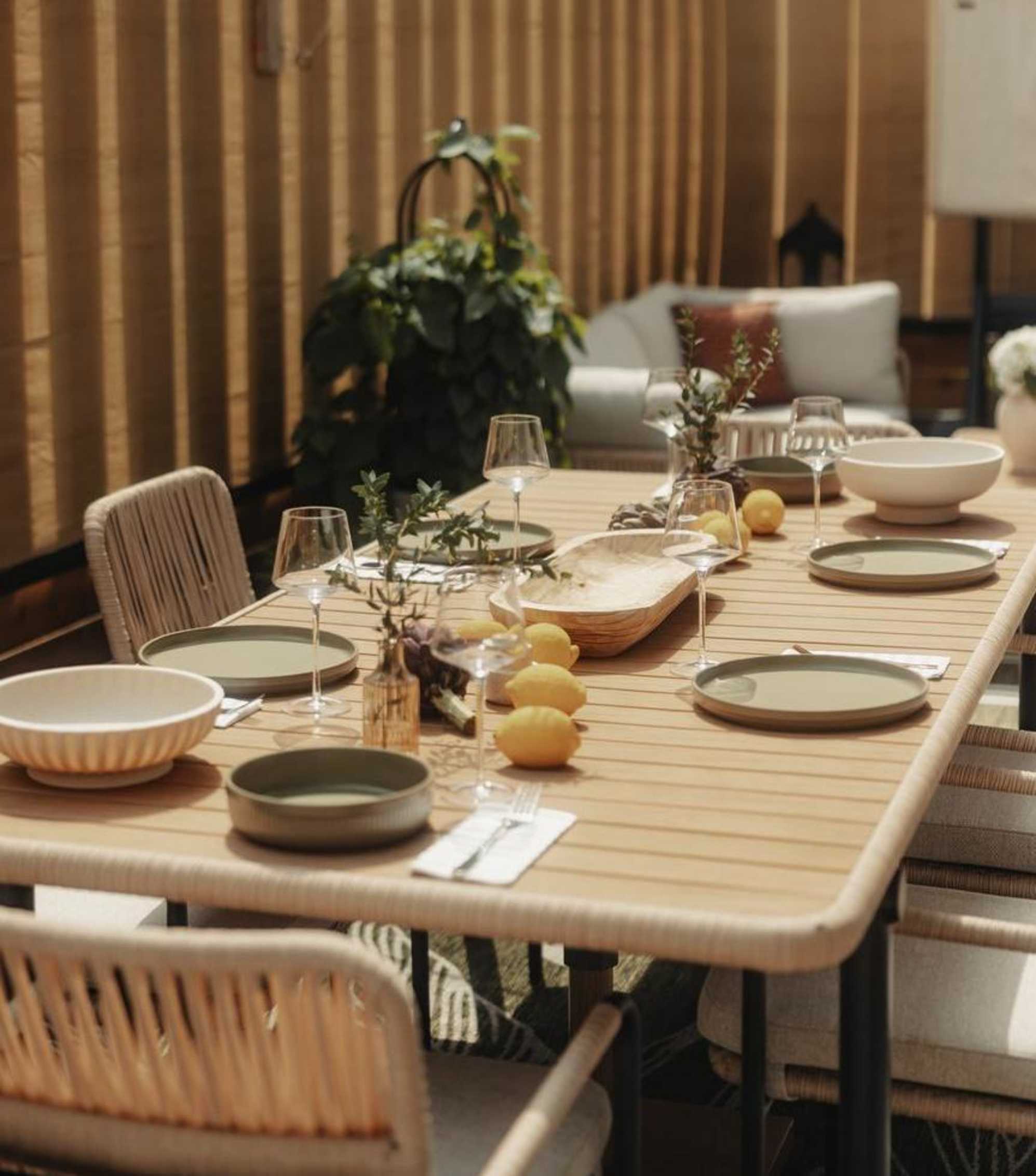 A wooden outdoor dining table with plates, bowls, and wine glasses placed on the tabletop.