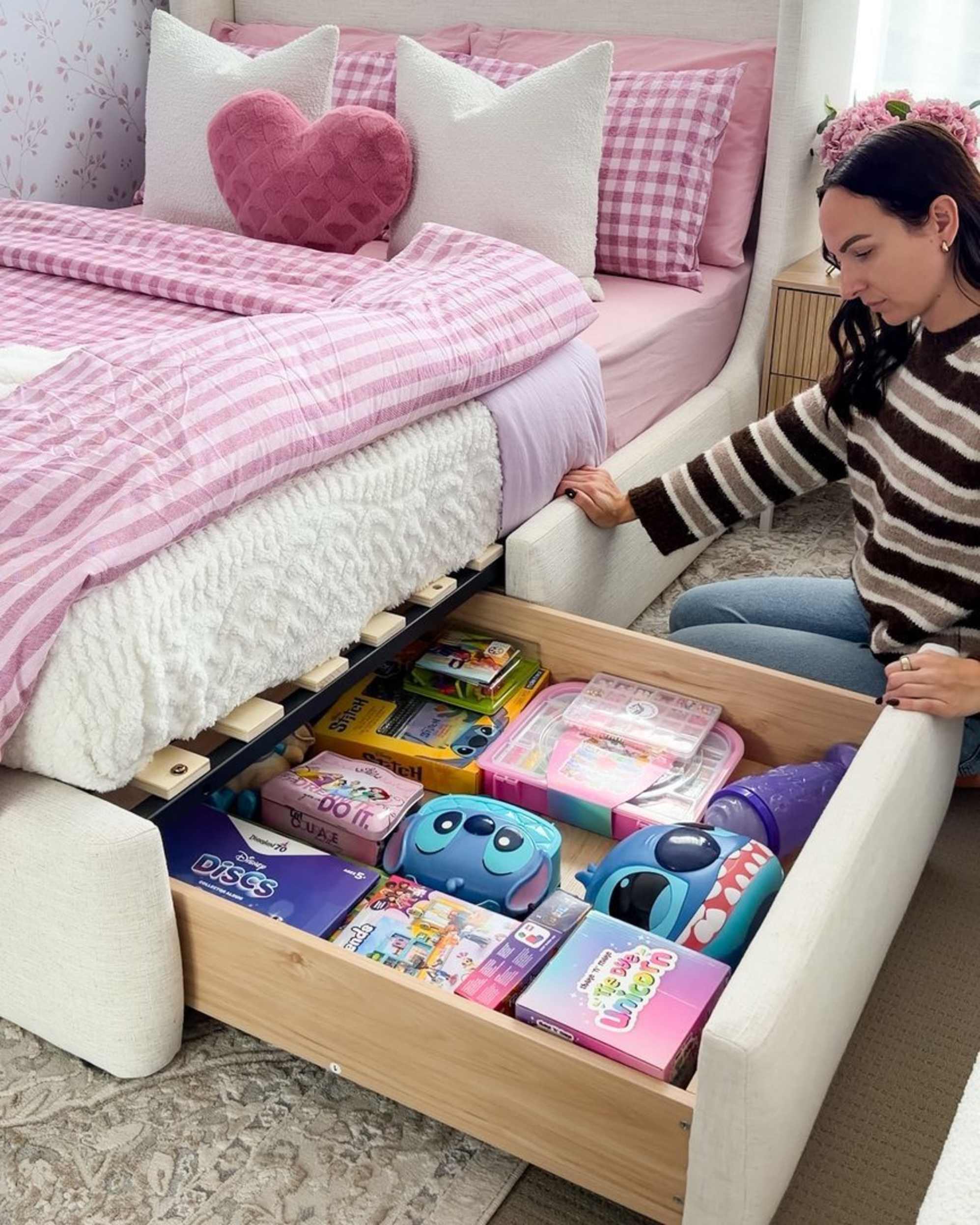 A woman kneeling on the bedroom floor as she pulls out a drawer from a storage bed to reveal children's toys.