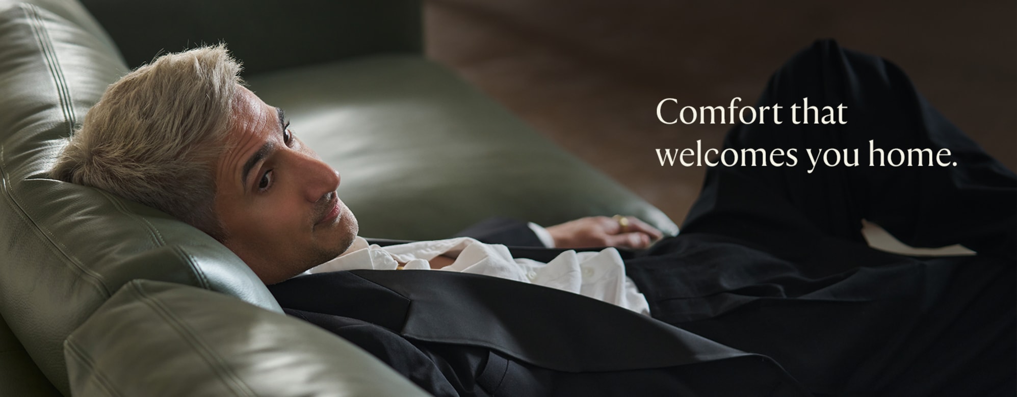 Tan France in a suit reclining on a green leather sofa.