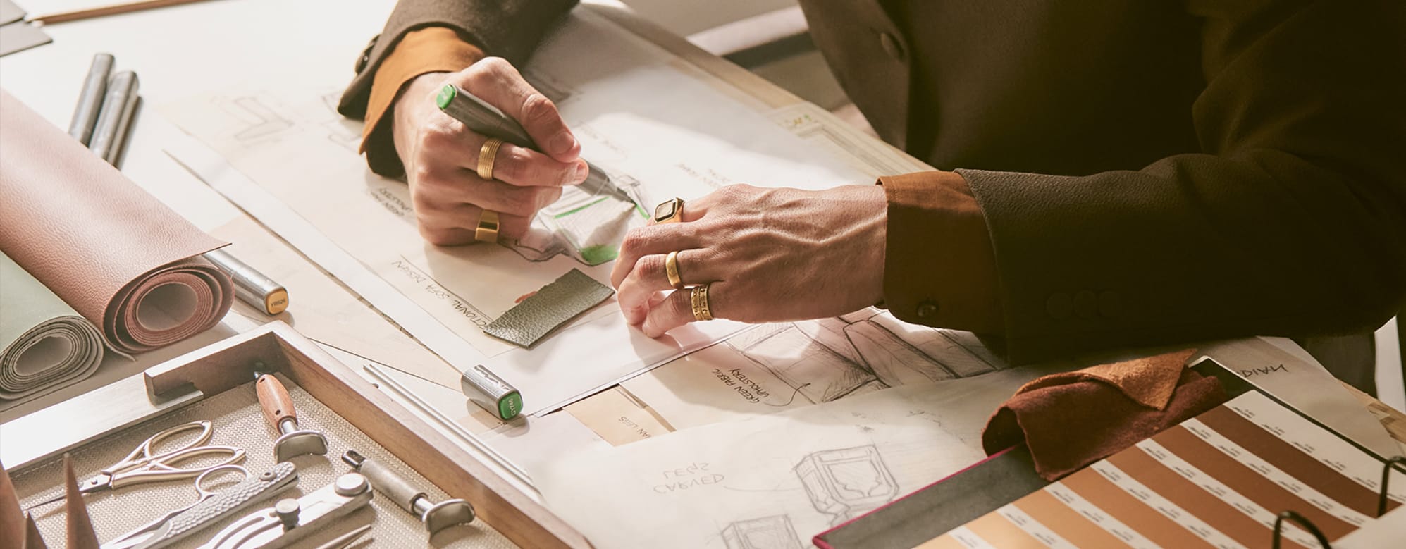 Close-up of a designer's hands sketching furniture upholstery details on paper, surrounded by leather swatches, fabric samples, and design tools on a studio desk.