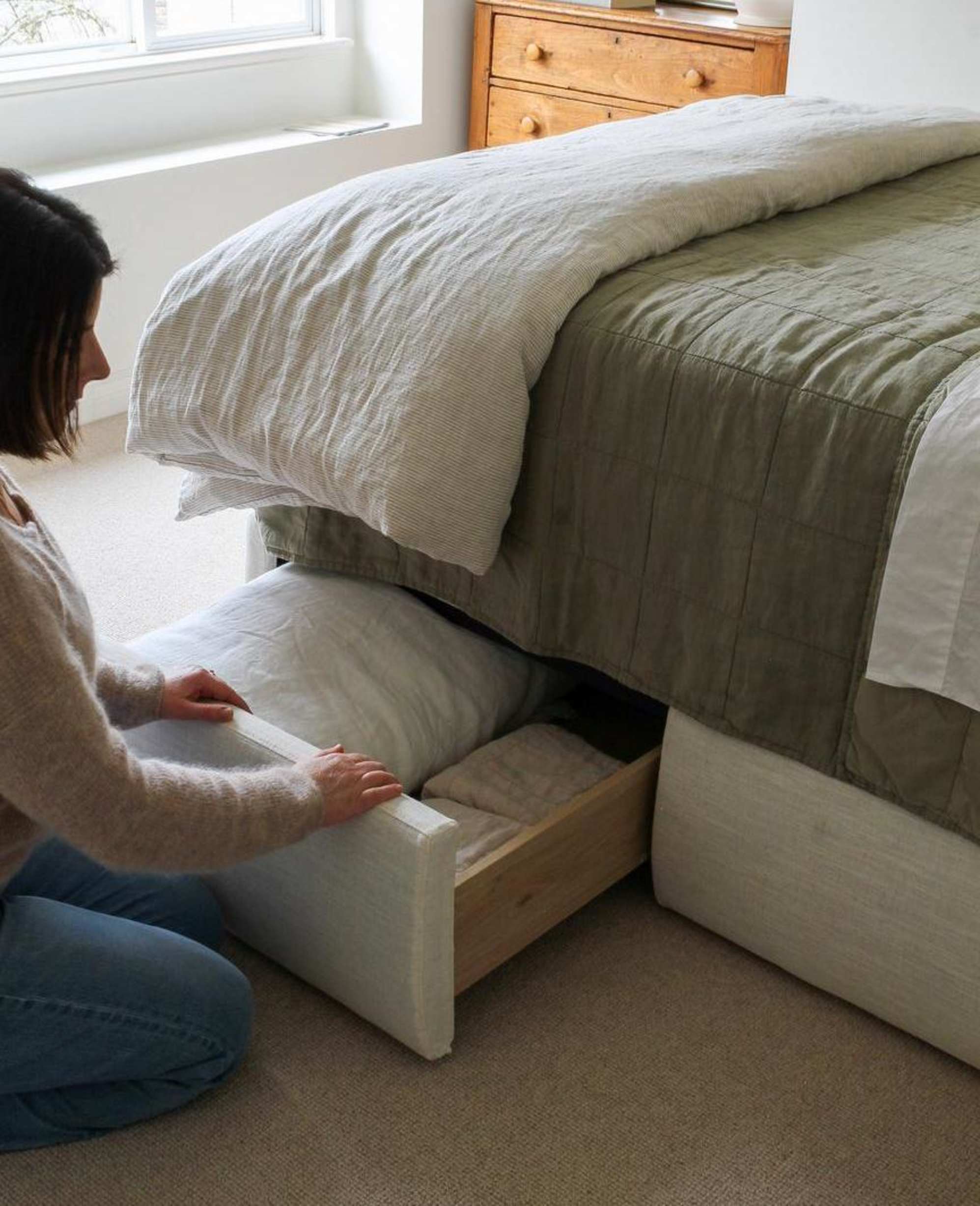 A person sitting on a floor while pulling open a drawer of a drawer storage bed to reveal bedding.