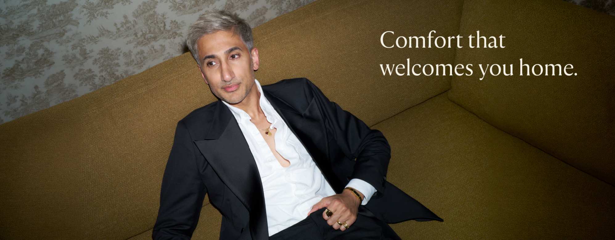 Tan France seated on a mustard-colored sofa in an open black suit.