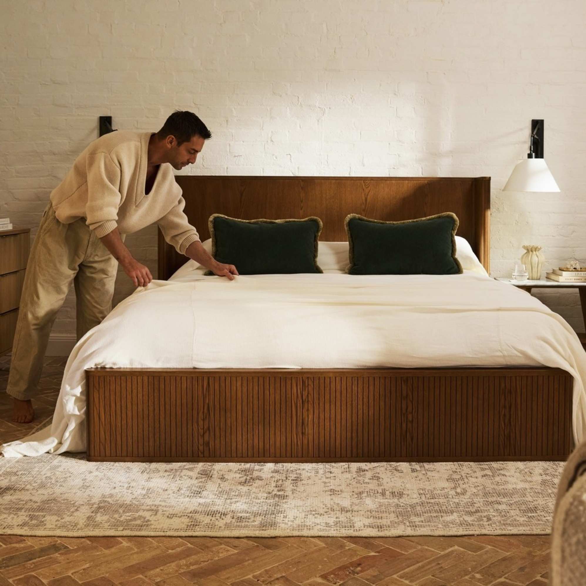 A man adjusting the bedsheets on a wooden bed with a wingback headboard.