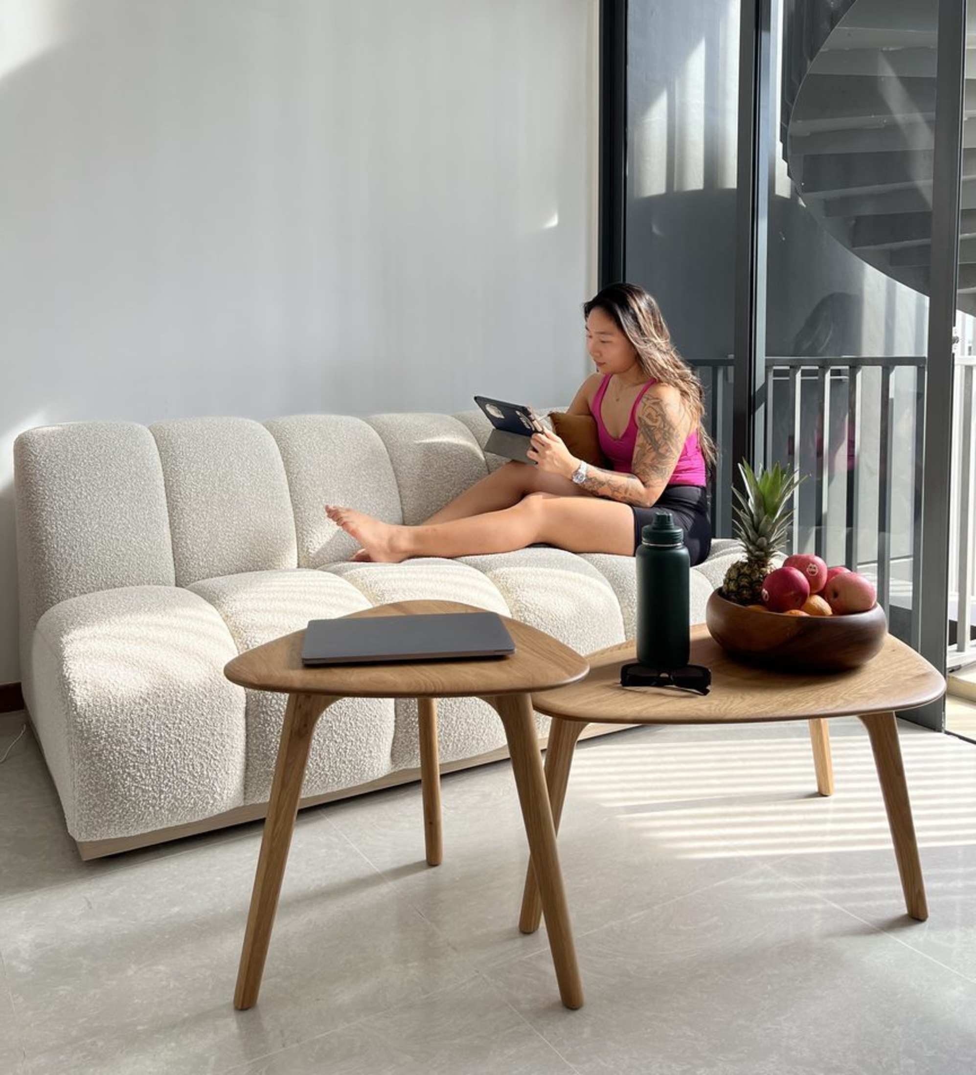 A woman sitting on an armless sofa while using her iPad, with two nesting coffee tables in front of her.