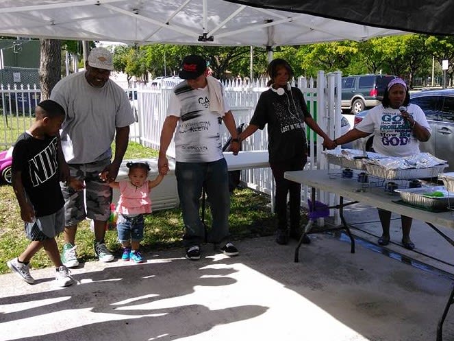 Mama Joy blesses the food at an event. Several guests hold hands with her.