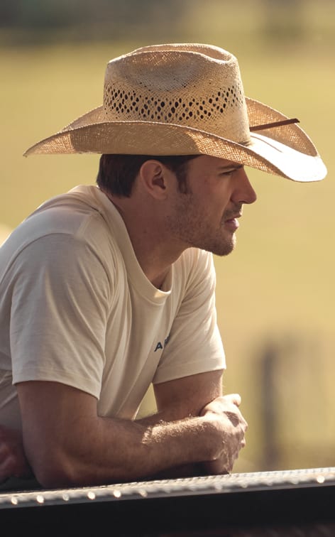 A man wearing a light-colored T-shirt and a straw cowboy hat with decorative cutouts, leaning on a railing and looking into the distance. The background is blurred, showing a warm, sunlit outdoor setting.