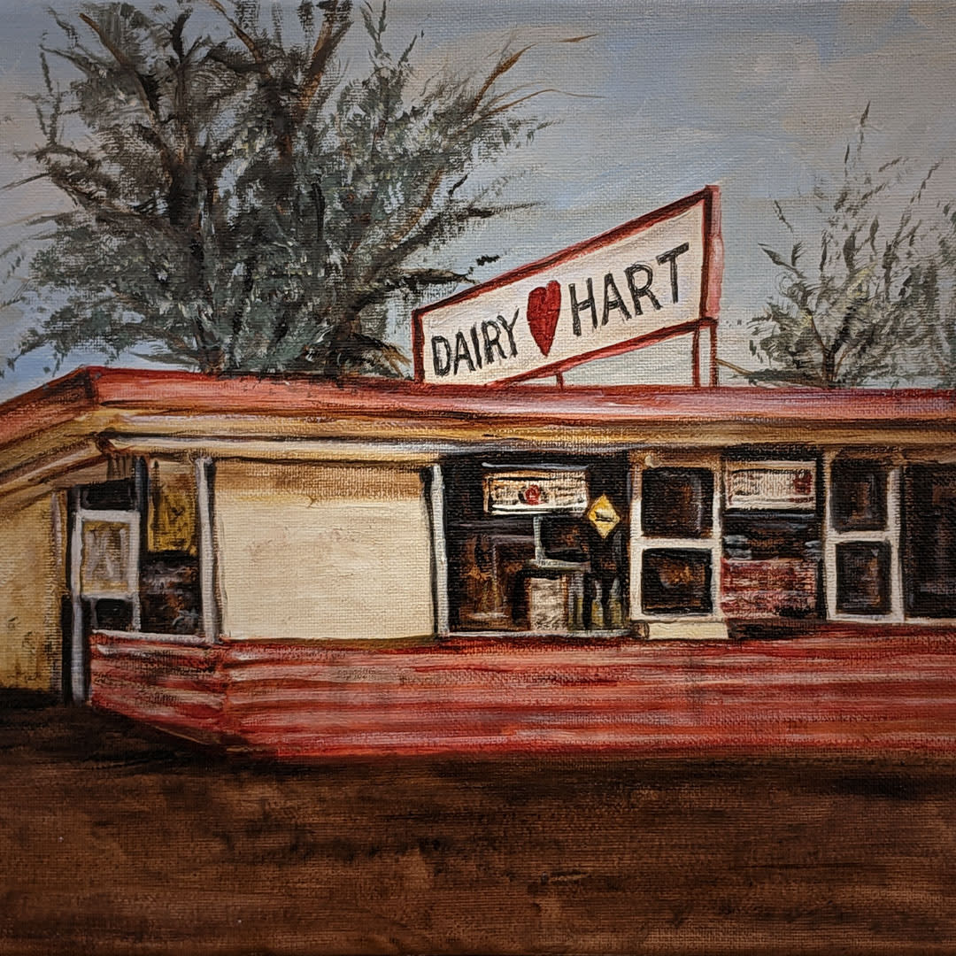 The image shows a nostalgic painting of a Dairy Hart, a quaint vintage-style diner from 1957. The building features a classic mid-century design with a prominent, slightly angled roof and a wide front that includes large windows and a central entrance. The signage above reads "Dairy ♥ Hart" in bold, red letters with a heart symbol, capturing a charming, retro feel. A large tree looms over the building, adding a touch of nature to the scene, depicted under an overcast sky that enhances the moody, historical ambiance of the painting.