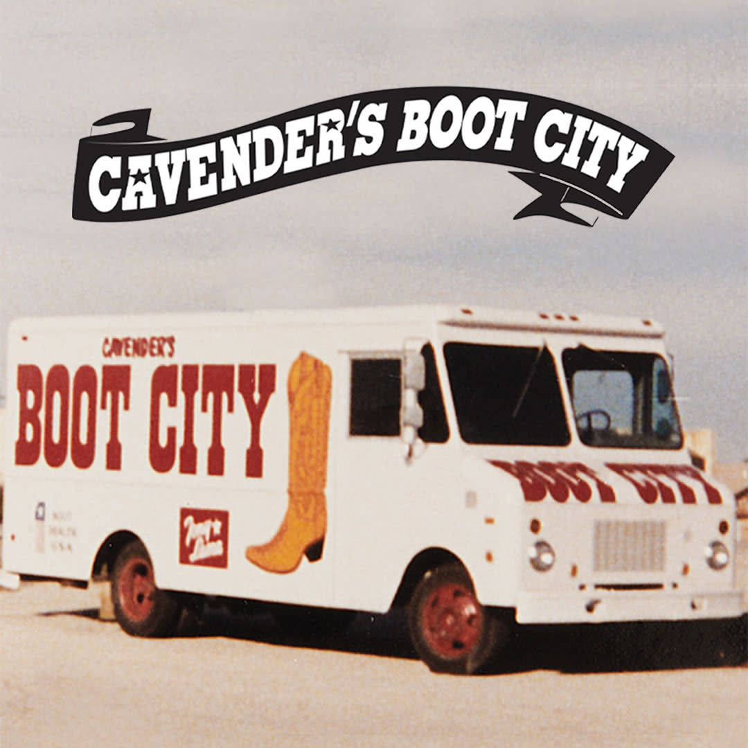 The image features a vintage photo of Cavender's Boot City delivery truck, capturing a classic piece of Americana. The truck, likely from the late 20th century, is painted white with large, eye-catching graphics that include a stylized image of a cowboy boot and the words "Cavender's Boot City" with a small Tony Lama logo along the side. The Cavender's logo, prominently displayed on the truck, uses a bold, Western-style font, adding to its thematic appeal. The background is a clear, expansive sky, suggesting the truck is situated in a spacious, possibly rural area. This nostalgic image highlights Cavender's long-standing presence in the Western apparel market.