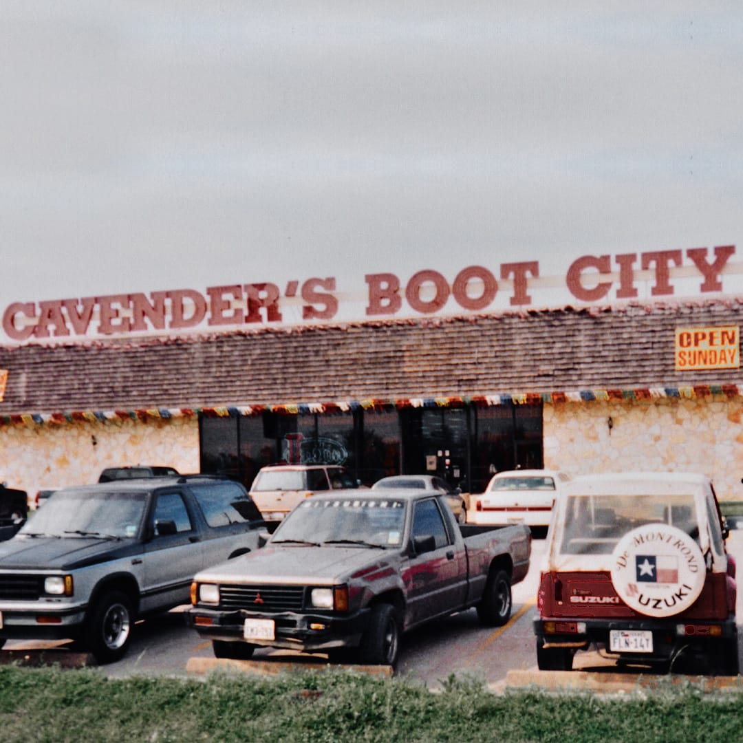 The image depicts a vintage photograph of Cavender's Boot City store, characterized by its rustic stone facade and a large, prominent red sign above featuring the store’s name. The parking lot in front is filled with cars from the 1980s, indicating the era. A sign stating "Open Sunday" is visible on the right, suggesting the store's flexible hours. This nostalgic scene captures a slice of American retail history, emphasizing the long-standing appeal of Cavender’s in the community.