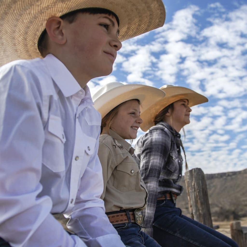 Three young children in cowboy attire with hats and boots, seated on a wooden fence, captivated by an outdoor western event.