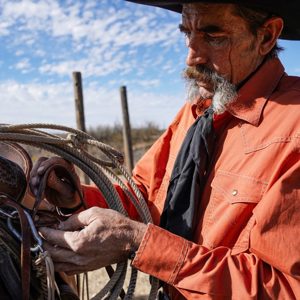 A seasoned cowboy with a distinguished black and gray mustache, donning a classic cowboy hat and an orange western shirt, securing his rope to the saddle in preparation for tending to cattle.