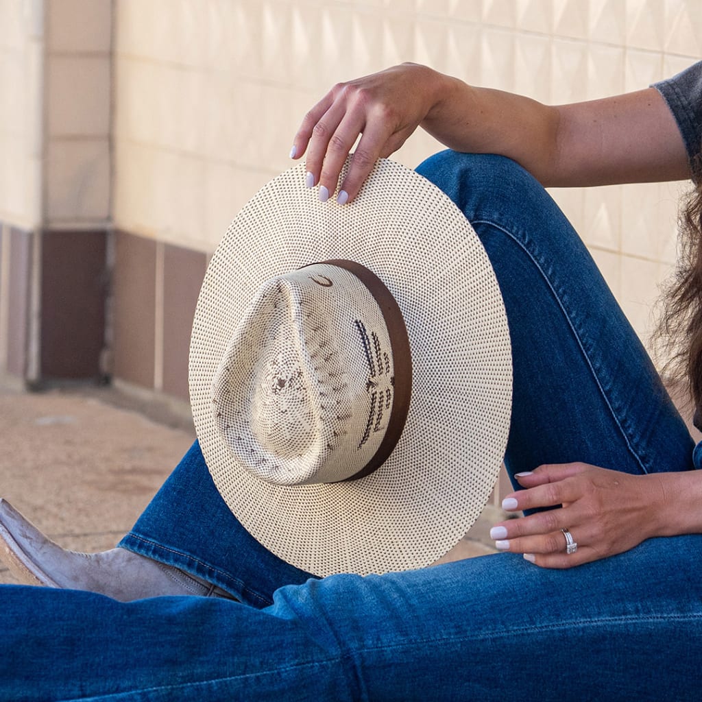 A young lady wearing blue jeans and cowgirl boots sitting on the ground holding a straw cowgirl hat on its side with brim facing forward.