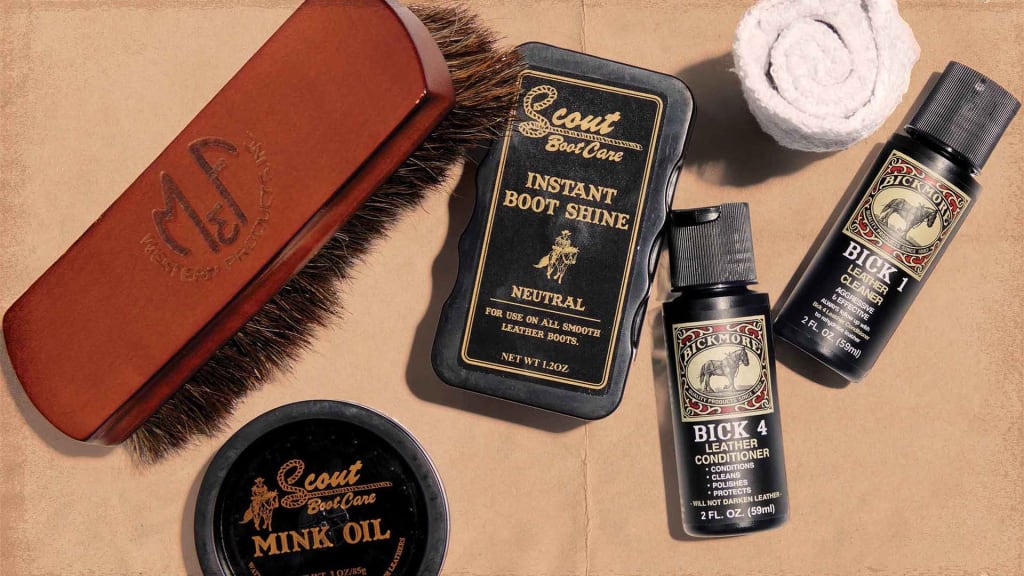 An assortment of cowboy boot care products arranged on a paper-like textured background. The items include a large brown horsehair brush with a logo etched into the wooden handle, a tin labeled 'Scout Boot Care Mink Oil' for conditioning, a bottle of 'Scout Boot Care Instant Boot Shine' in a black container with a neutral color indication, a small white rolled-up cloth, and two bottles of 'Bick 4 Leather Conditioner' which clean, condition, polish, and protect leather. These products suggest a comprehensive boot maintenance kit.
