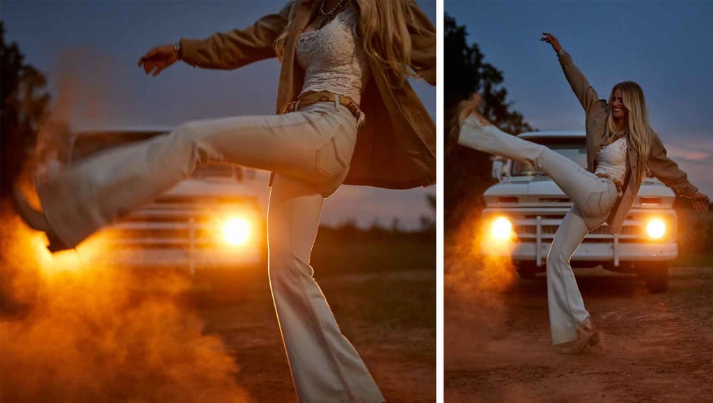Ariat Women's Denim Jeans Fashion Moment: with a happy cowgirl girl kicking her boots in a dirt parking lot with a classic pickup truck with it headlights on at dusk,