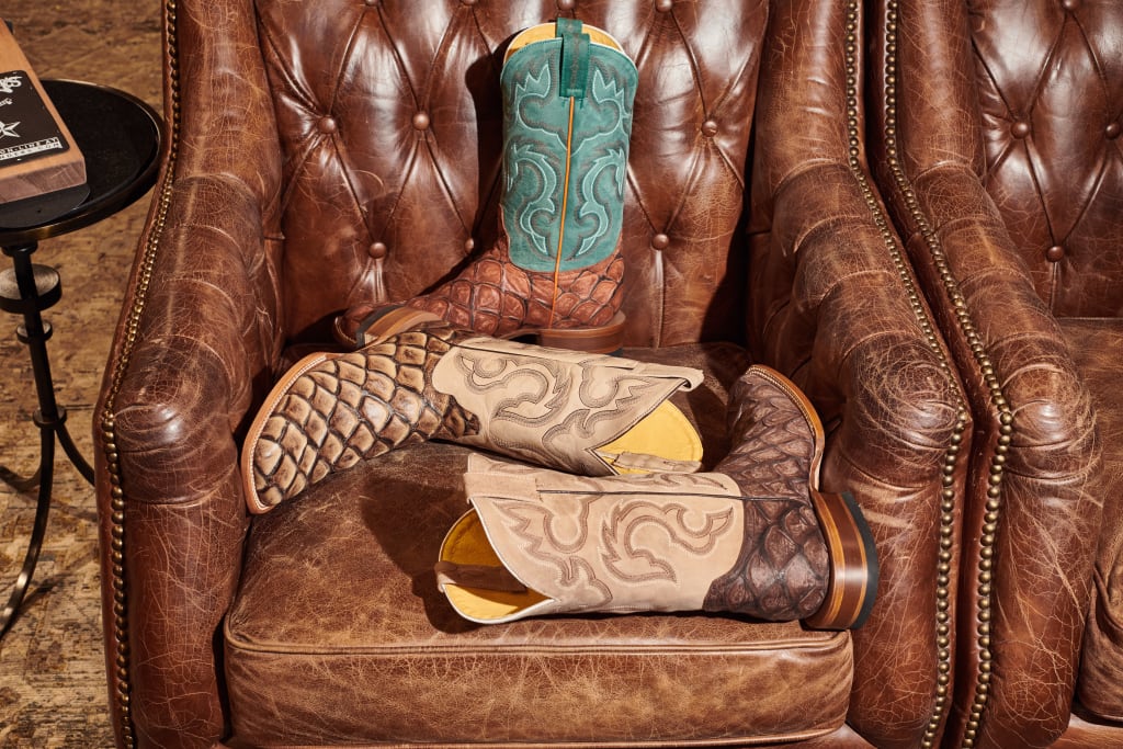 Image of three exotic cowboy boots resting on a brown distressed leather armchair with nailhead trim and button tufting. Each boot features a fish-scale construction in varying colors of tan, brown, cognac, and teal.