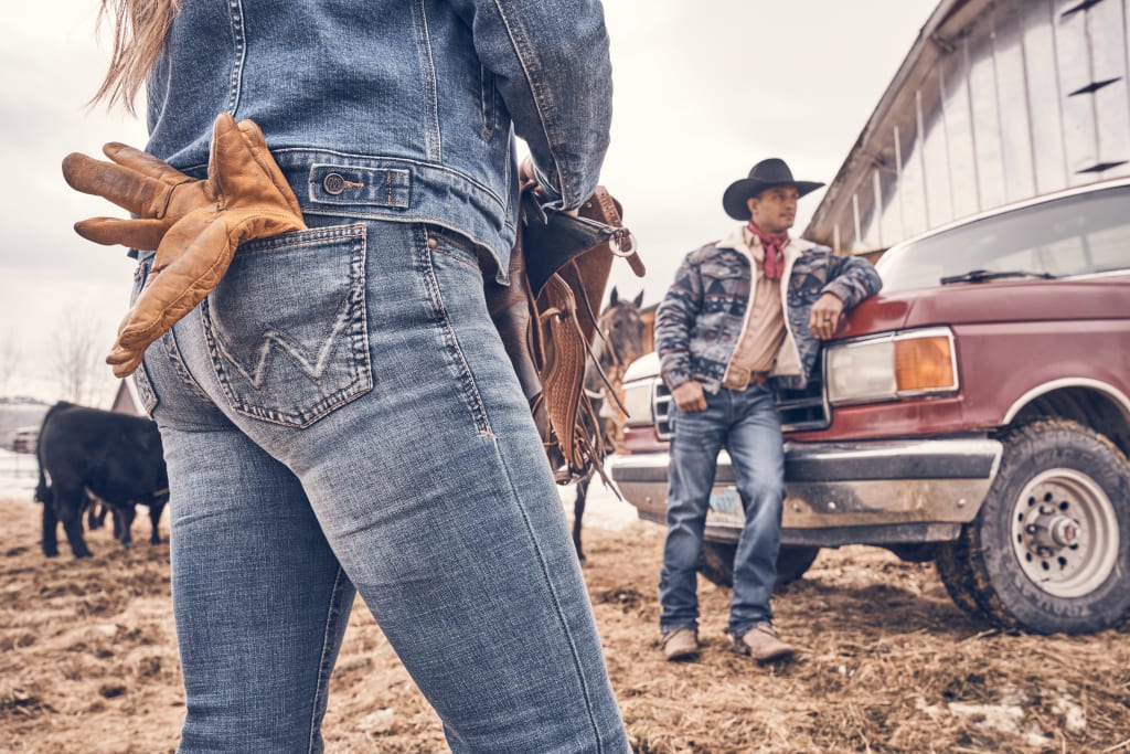 A cowboy leaning on a Ford pickup truck, accompanied by a cowgirl wearing jeans with work gloves in back pocket, set against a rustic farm backdrop. A black cow stands on the left side of the photo, with a dirt ground completing the scene.