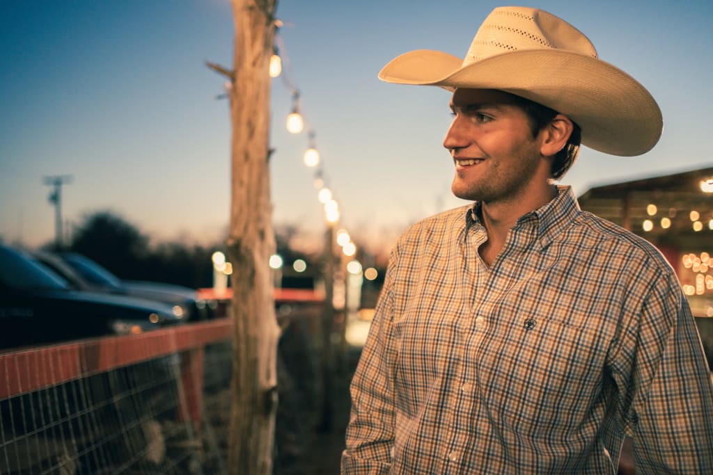 Cowboy wearing a classic western hat, smiling, gazing towards a parking lot illuminated by decorative evening lights at an outdoor event.