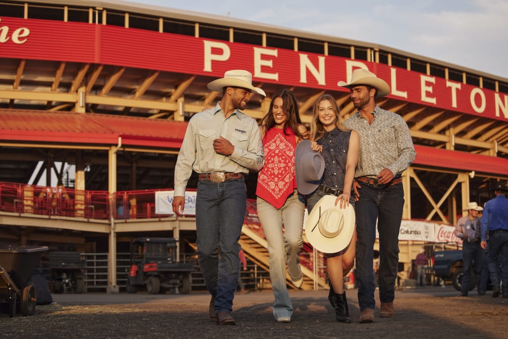 Two couples in coordinated western wear, walking closely with beaming smiles. They don stylish cowboy hats, jeans, and boots, against the backdrop of a sunset at a rodeo or country event.